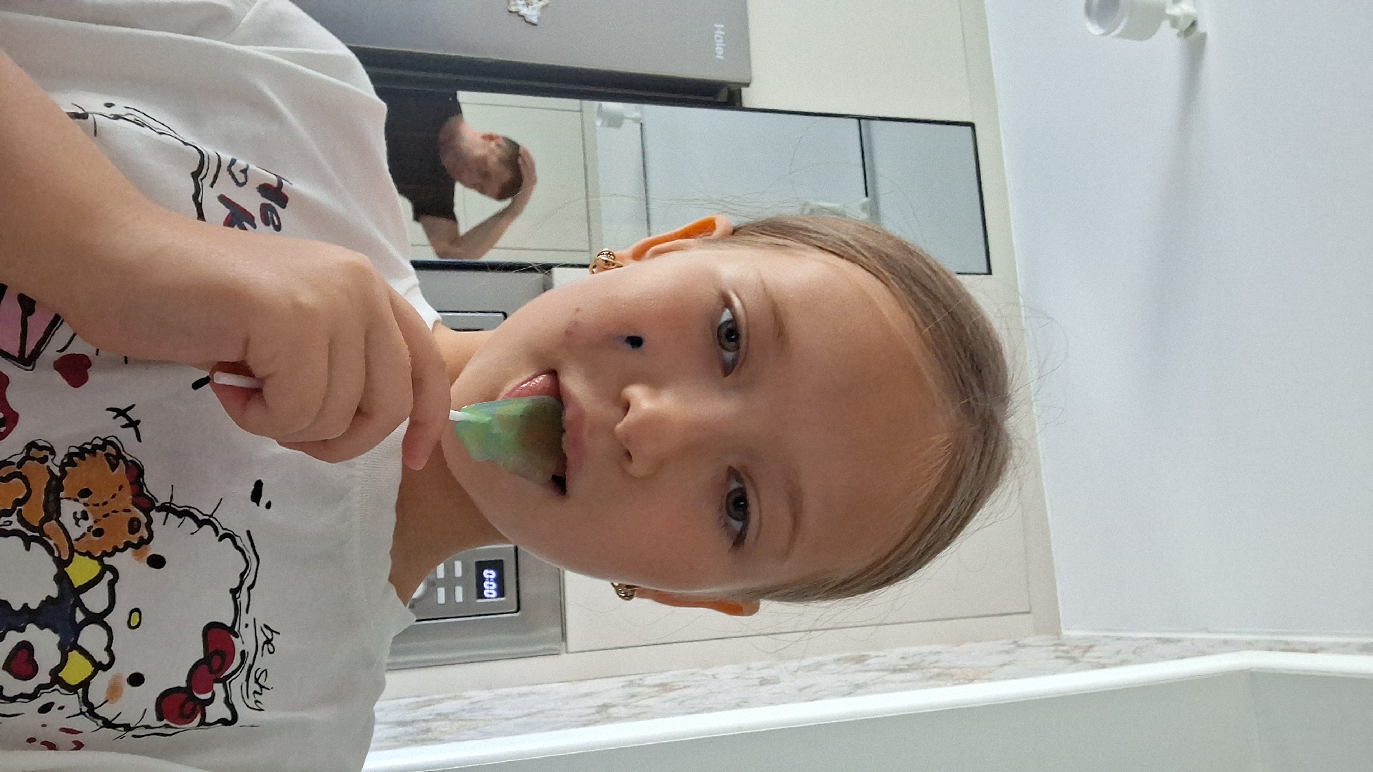 The image shows a young girl with blonde hair and light skin, looking at the camera with her mouth open, holding a green popsicle. She is wearing a white t-shirt with a cartoon design featuring Hello Kitty and a teddy bear. In the background, a man is visible in a mirror, with his hand on his head, looking down. There is a kitchen appliance with a digital display that shows "0:00". The setting appears to be a kitchen or bathroom, with white cabinets and a countertop. The lighting suggests it is daytime. The girl's expression is playful.