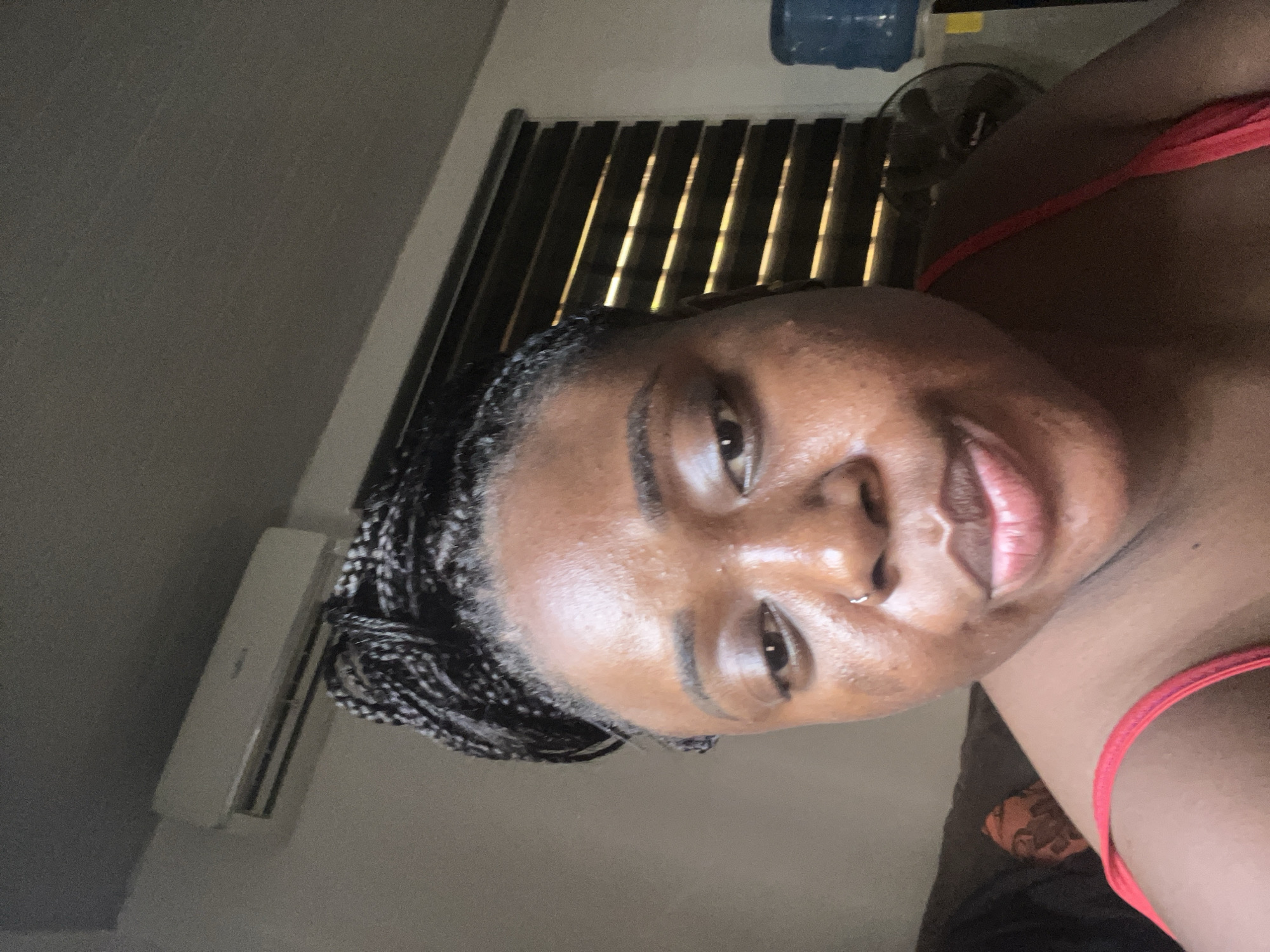 The photograph shows a woman with dark skin and braided hair wearing a red top. She has a nose piercing on her left nostril. The background features closed window blinds, a blue water dispenser, a fan, and an air conditioning unit. The image appears to have been taken indoors. There is no information provided to assume the location is in the city of Abuja, Nigeria.