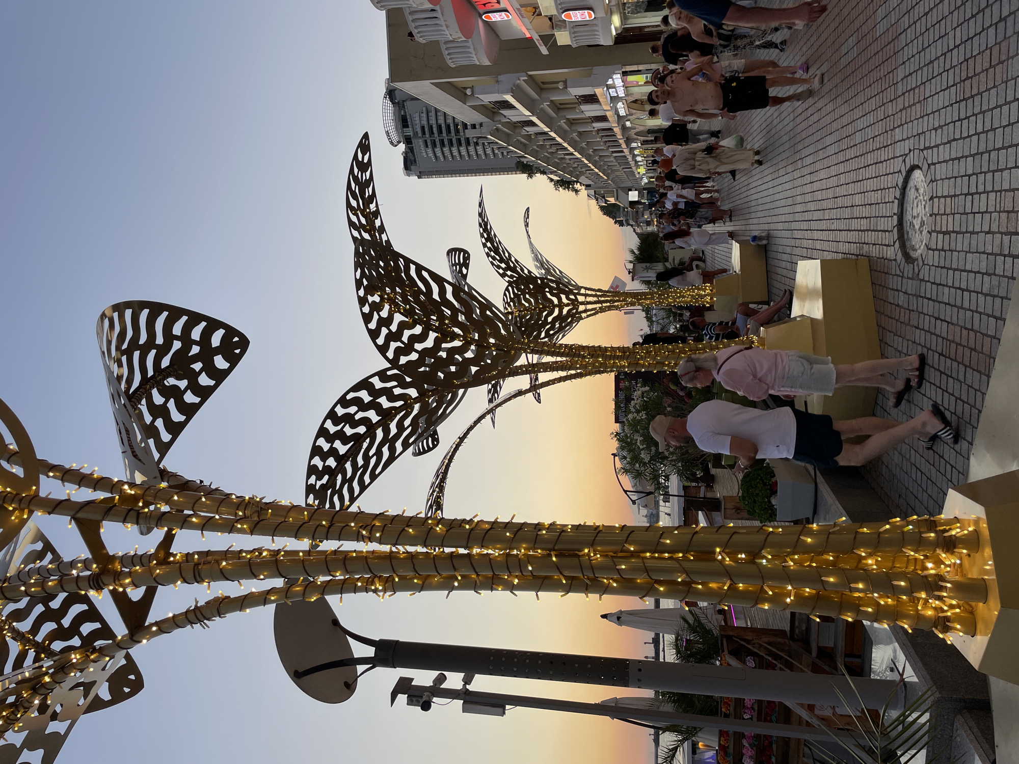 The image depicts an evening scene along a bustling pedestrian walkway in Sochi, Russia, likely on the embankment. In the foreground, decorative palm tree-like structures wrapped in warm-toned fairy lights stand prominently. The sky shows a gradient of sunset colors, transitioning to a clear dusk blue.
A diverse crowd of people are seen strolling along the paved walkway. They are dressed in casual summer attire, suggesting a warm climate and a leisurely atmosphere. Some individuals are walking towards the viewer, while others are walking away. There are shops and restaurants lining the right side of the walkway, with some displaying illuminated signage, including a Burger King.
Further in the background, a tall modern building rises above the lower structures, indicating an urban setting. The left side of the walkway features some outdoor seating areas, greenery, and umbrellas, hinting at cafes or beach access. The overall impression is one of a vibrant tourist area enjoying the transition from day to night.