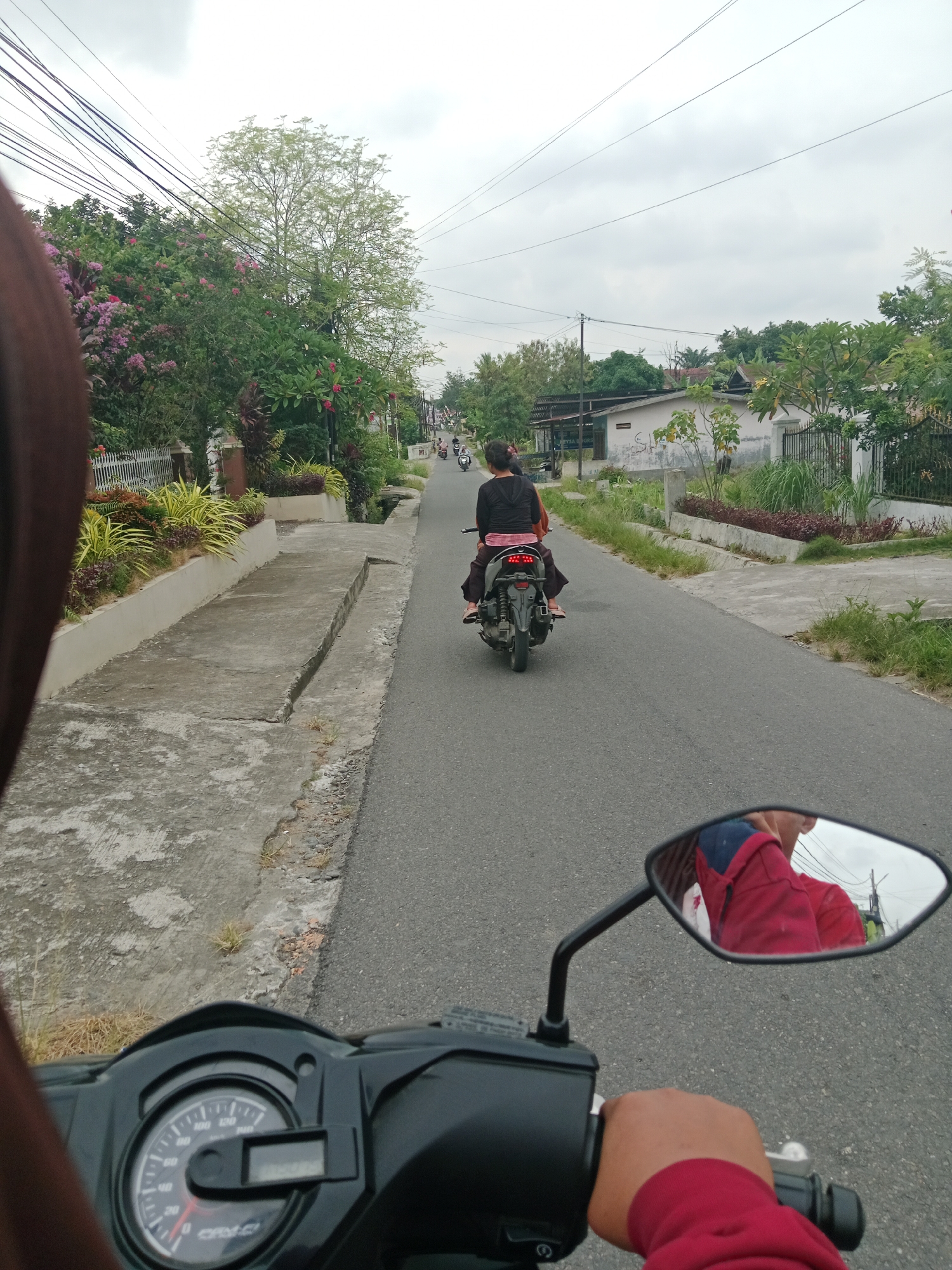 The image captures a first-person perspective of someone riding a motorcycle on a paved road in the city of Binjai, Indonesia. In the foreground, the motorcycle's handlebars and a portion of the instrument panel are visible. A side mirror reflects a person wearing a red jacket. The road is lined with a concrete sidewalk and vegetation, including trees and flowering plants. Further down the road, another motorcycle is seen carrying two passengers. Houses and more greenery are visible on the sides of the road. The sky is overcast, suggesting a cloudy day. The overall scene depicts a typical street view in a residential area of Binjai.