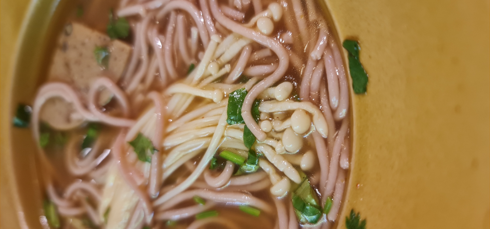 A close-up, high-angle shot depicts a bowl of Vietnamese noodle soup, potentially Mỳ Quảng or a similar dish, served in a light yellow bowl. The soup broth is a clear brown, and the primary ingredients are noodles (predominantly a light pink hue), white enoki mushrooms, squares of what appears to be pork sausage or a similar processed meat, and green herbs, likely coriander or scallions, used as a garnish. The soup is positioned off-center, taking up the majority of the frame. There are no people visible. The setting or location cannot be definitively determined from the image alone, though the presence of Vietnamese cuisine would suggest the location might be in Vietnam, possibly Đà Nẵng as requested in the prompt, though without further information, this is speculative. The overall composition emphasizes the textures and colors of the ingredients within the soup.