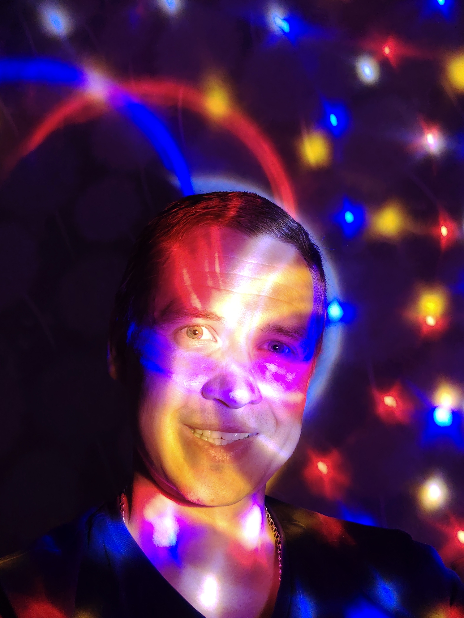 A medium shot captures a man indoors, illuminated by a vibrant array of colored lights. The lights, consisting of red, blue, and yellow hues, cast a dynamic pattern across his face and the background. The man, facing the camera, is smiling, with one eye appearing lighter than the other due to the light pattern. He is wearing a dark shirt and a gold chain is visible around his neck. The background is filled with scattered, out-of-focus lights, creating a festive atmosphere. The image location is not discernible as Chaikovsky, Russia from the visual information provided.