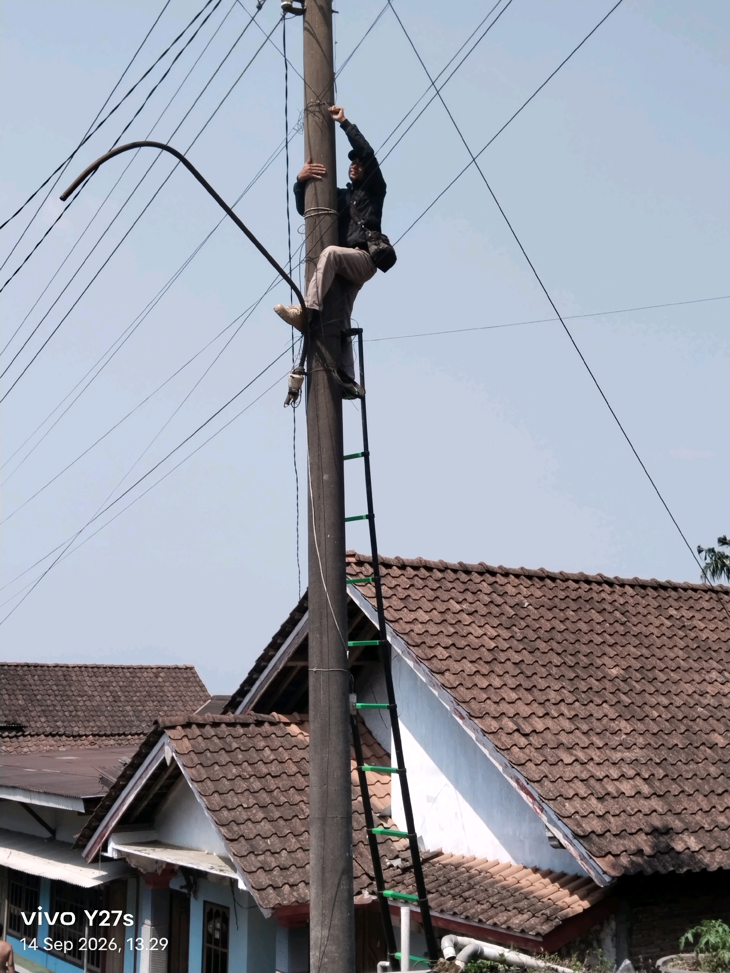 An individual wearing a dark jacket, light-colored pants, and a dark cap is positioned on a utility pole in Tamansari, Indonesia. The person is holding onto the pole and appears to be working with cables. A ladder is attached to the pole, extending upwards. Wires are strung across the sky, and residential buildings with tiled roofs are visible below the pole. The sky is clear and blue. The timestamp "14 Sep 2026, 13:29" and "vivo Y27s" are superimposed on the lower-left corner of the image.
