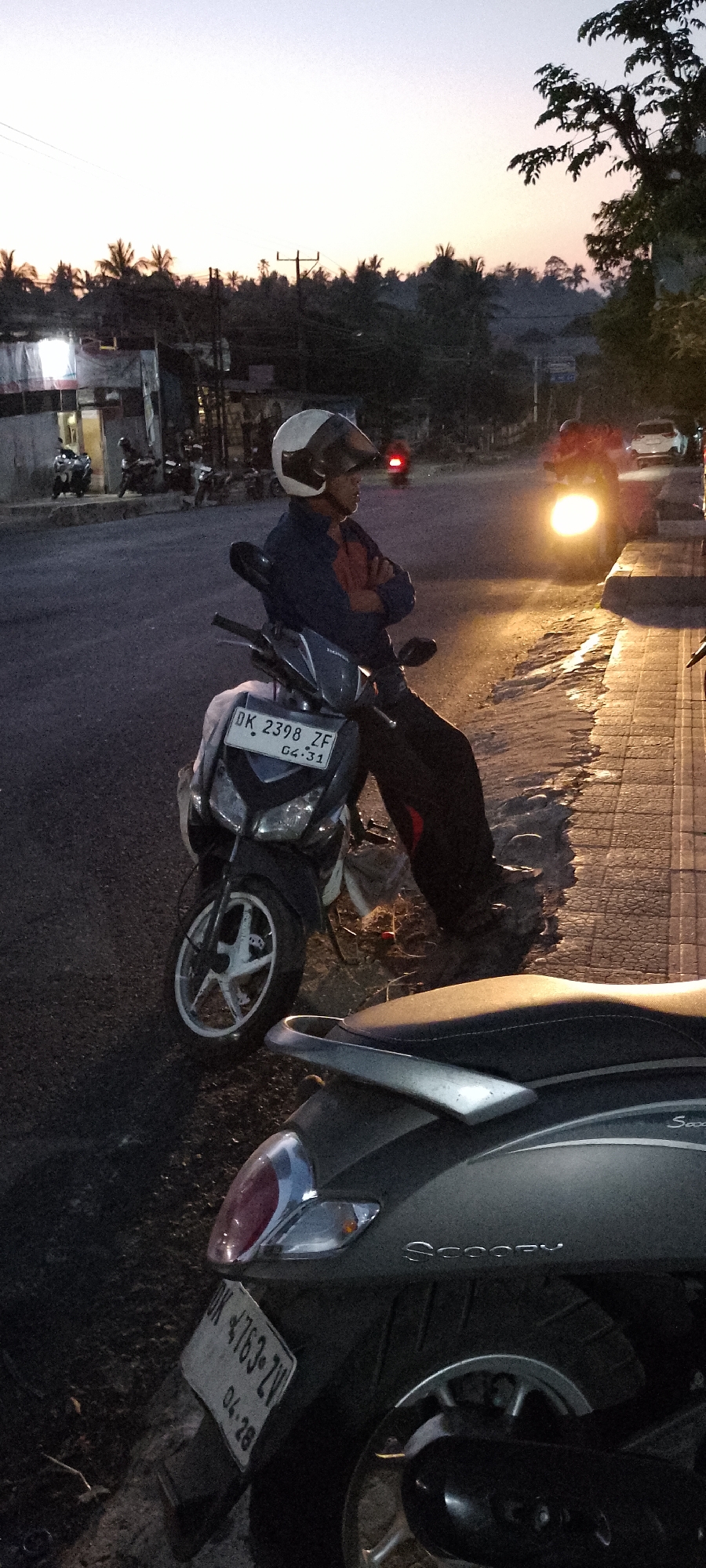 The image depicts a street scene in Pekutatan, Indonesia, during twilight. A man wearing a white and black helmet and a dark blue jacket is seated on a dark-colored scooter. He is looking towards the right side of the frame with his arms crossed. The scooter has a license plate with the characters "DK 2398 ZF 04.31". Another scooter is partially visible in the foreground, with its license plate showing "DK 4763-ZV 04.28". Several other motorcycles are parked in the background. The street is paved, and a sidewalk with visible tiles is on the right. Buildings with lights are present on the left side of the street, and palm trees and other vegetation form a dark silhouette against the pale twilight sky. The headlights of a vehicle are illuminated in the distance.