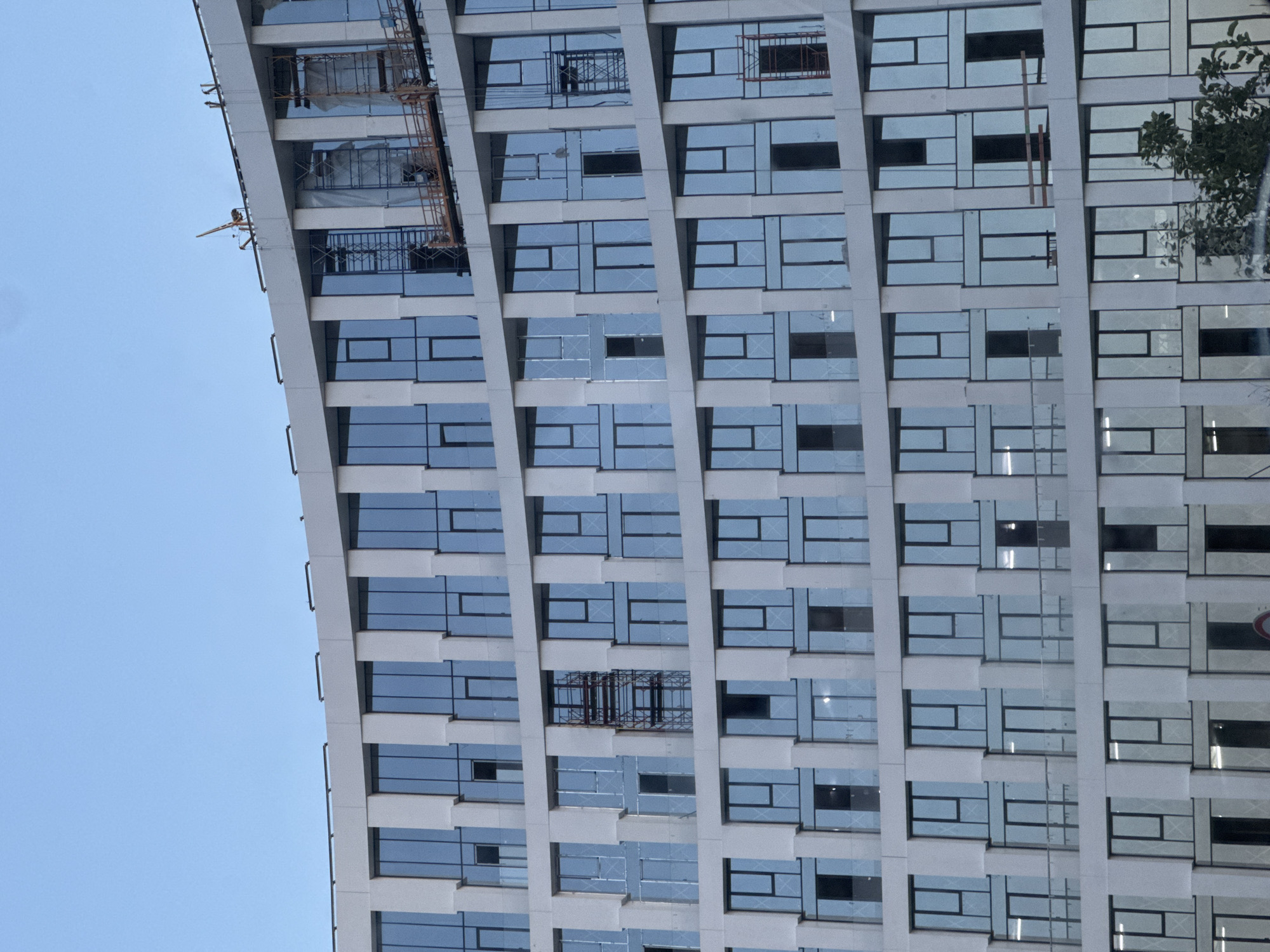 A multi-story, modern building with a curved facade, currently under construction, is depicted against a clear, light blue sky in the city of Unknown, Unknown. The structure features a grid pattern of rectangular windows framed by light-colored, possibly concrete or stone, vertical and horizontal panels. The windows predominantly reflect the blue sky, with some showing interior scaffolding or protective coverings.
On the upper levels, evidence of ongoing construction is visible: a small yellow tower crane is positioned on the rooftop towards the right, and a suspended work platform with a wooden deck and metal railing hangs from the upper facade. Multiple sections of blue and red metal scaffolding are visible within window openings on various floors, with some interior sections covered by white tarpaulin. The building appears to have at least seven visible floors. The lighting suggests daylight, with soft illumination and no harsh shadows. A small cluster of dark green foliage partially obscures the building's bottom right corner. No human figures are discernible in the frame.