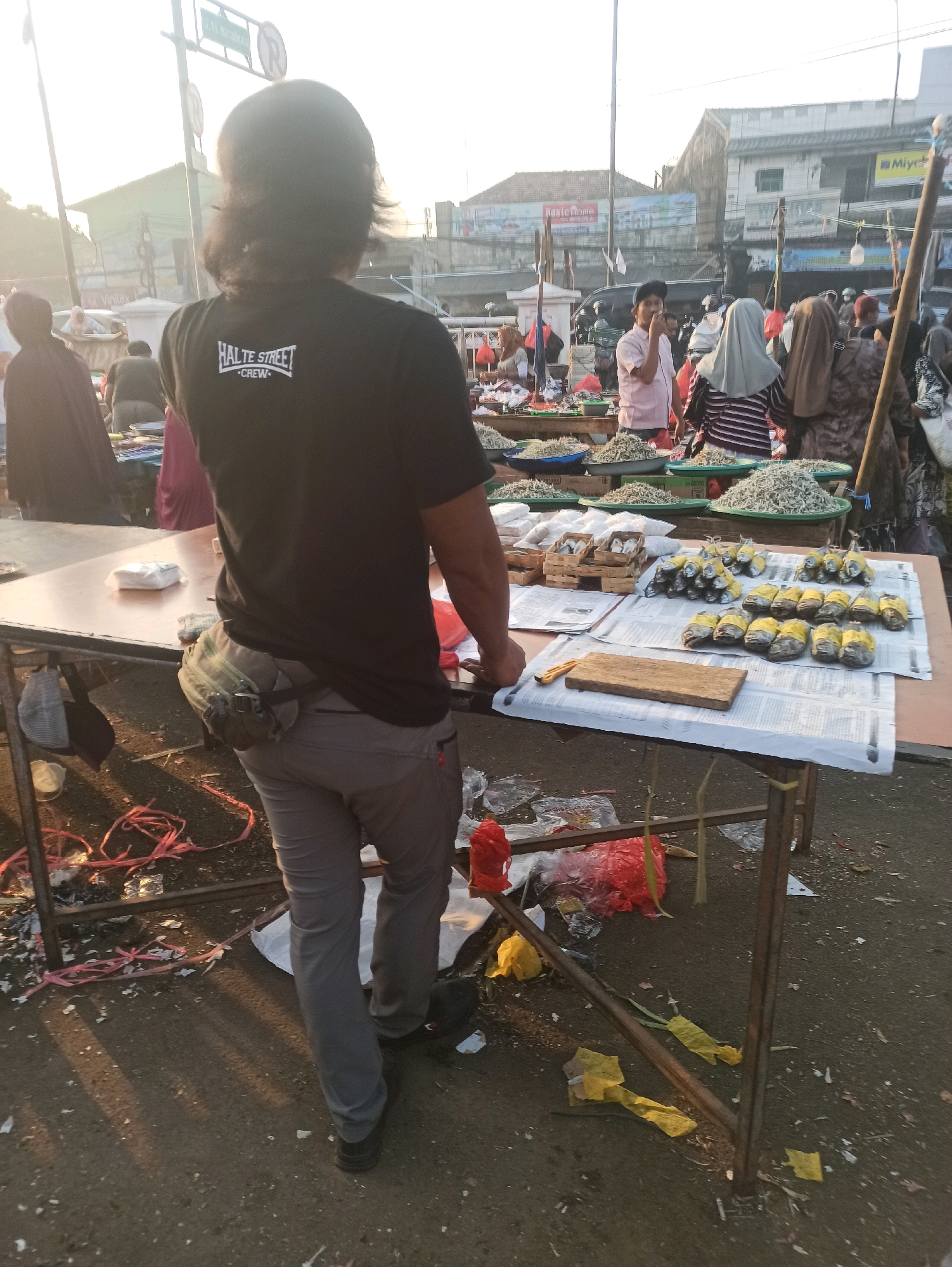 The image depicts an outdoor market scene in Cikarang, Indonesia. In the foreground, a person wearing a black t-shirt with "HALTE STREET - CREW" printed on the back is standing with their back to the camera. They are wearing grey pants and a fanny pack around their waist. The person is standing next to a market stall covered with a table displaying various items.
The table is laden with goods, including what appear to be small fish or anchovies in plastic containers and bowls. There are also some wrapped food items, possibly fish, wrapped in yellow material. A small wooden cutting board and a knife are also visible on the table.
Behind the foreground stall, other market stalls and a crowd of people are visible. Several individuals are wearing headscarves, suggesting a predominantly Muslim population. The market appears to be set up outdoors with a dusty ground. In the background, buildings and street signs can be seen, indicating an urban or semi-urban environment. The sunlight suggests it is daytime, possibly early morning or late afternoon given the long shadows. There is a general sense of a busy marketplace with goods being sold.