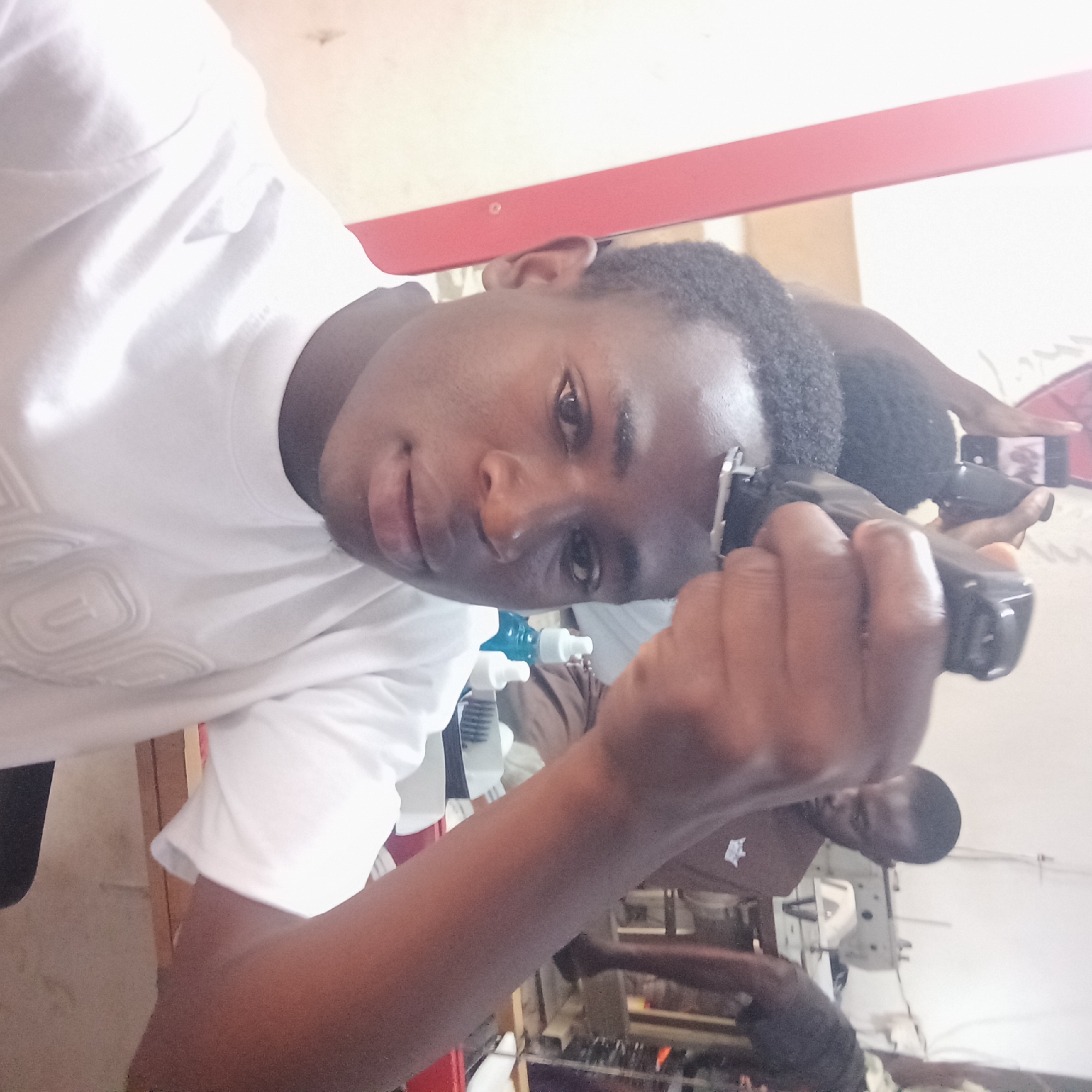 A young man is shown at close range, holding a black electric hair clipper to his forehead. He is light-skinned, wearing a white t-shirt, and looking directly at the camera. The clipper is in his right hand, angled towards the camera. In the background, a mirror reflects a partial view of the scene, showing another person holding a mobile phone. The location appears to be an indoor setting, possibly a barber shop, with various objects and equipment visible in the reflection. It is not possible to confirm the location as Jos, Nigeria, based on the visual information provided.