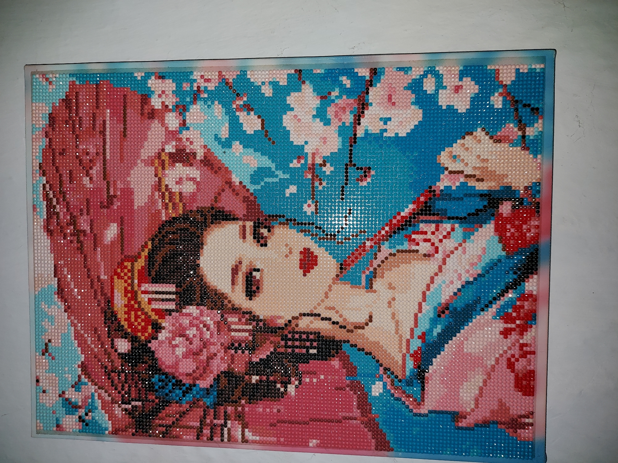 This is a close-up, well-lit, eye-level, vertical shot of a diamond painting of a geisha holding a parasol, set against a background of cherry blossoms. The painting is square-shaped and framed. The geisha is depicted with fair skin, dark hair styled with a pink flower and hairpins, and wears a blue and pink kimono. Her eyes are dark, and her lips are red. She holds a red parasol over her head. The background is a vibrant blue, with pink and white cherry blossoms scattered across it, with some branches visible. The diamond painting technique gives the image a textured, pixelated appearance. The overall color palette is bright and colorful, dominated by blues, pinks, and reds. The lighting is even, highlighting the facets of the diamonds. The background of the scene appears to be a plain white wall.