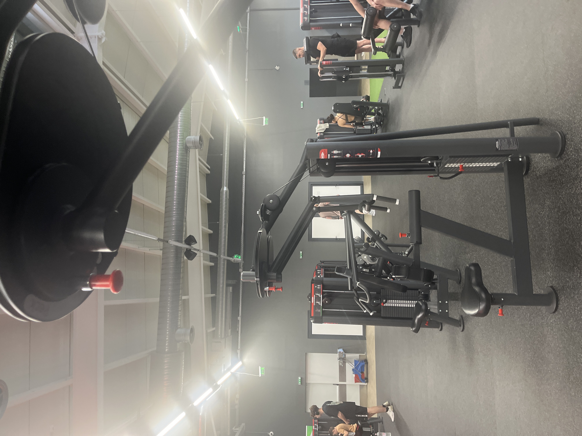 The image shows a gym interior with various exercise machines. The prominent machine in the foreground is a seated chest press. Other machines can be seen in the background with people using them. The gym has a dark gray floor and gray walls. There are white air ducts and lights on the ceiling. The image provides no information that places it specifically in Plovdiv, Bulgaria.
