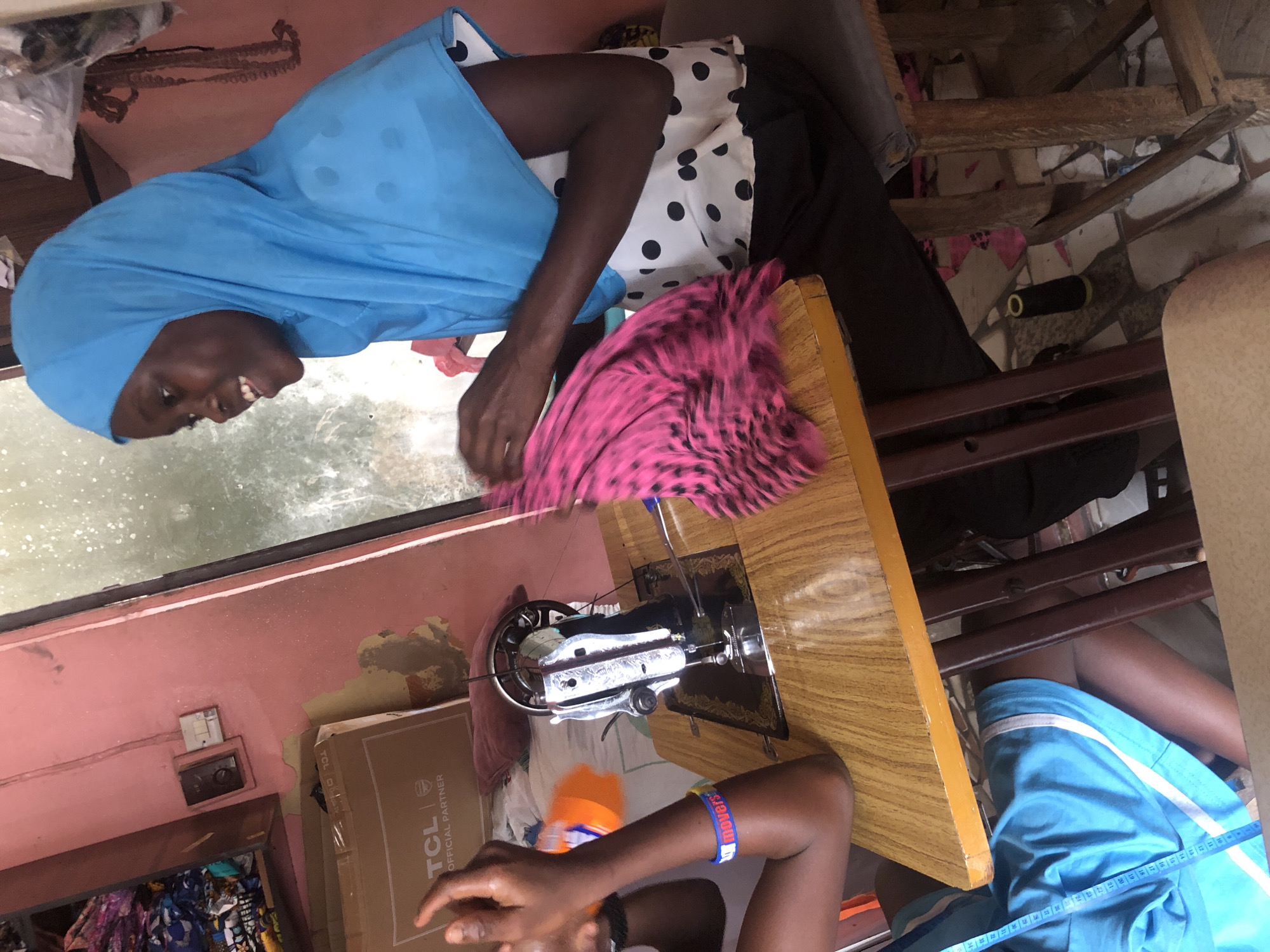 The image depicts a tailor's workshop in Ado, Nigeria. A young woman wearing a blue hijab and a white and black polka dot shirt is seated at a wooden table, actively engaged in sewing a vibrant pink and black patterned fabric with a vintage sewing machine. To her left, another person's arm is visible, holding an orange bottle and wearing a blue and yellow bracelet. The workshop environment is evident with a shelf of fabrics on the left, a TCL branded box, and a pink painted wall. The floor appears to be tiled. The scene conveys a sense of focused work and daily activity within a community tailoring space.