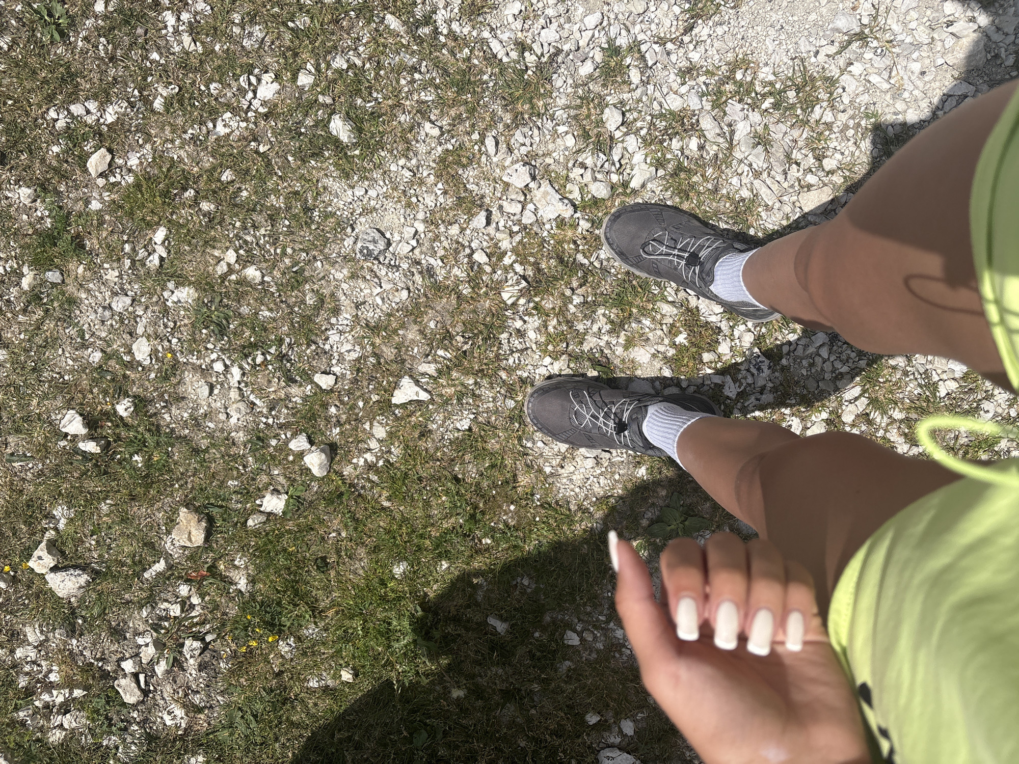 The image is a first-person perspective shot of a person's legs and hand, standing on a path covered in gravel and patches of grass. The person is wearing dark grey athletic shoes with white laces and white socks, and bright lime green shorts. A hand with white-painted fingernails is visible in the foreground, reaching towards the bottom right of the frame. The scene suggests an outdoor activity, likely walking or hiking, given the footwear and terrain. The lighting indicates it's daytime, with clear shadows cast on the ground, suggesting bright sunlight. The ground composition of small white stones and sparse green grass is consistent with a dry, possibly arid or mountainous environment. There is no visible text in the image.