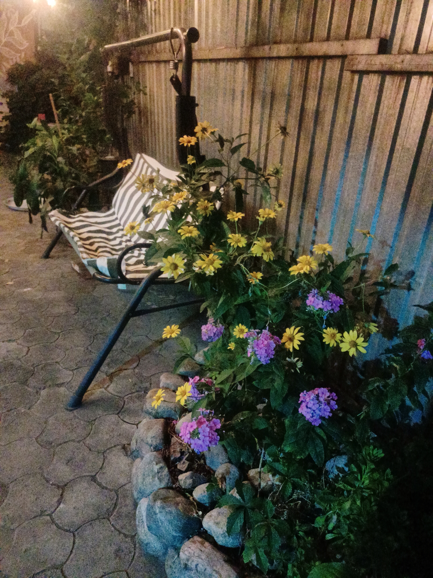 The image depicts an outdoor scene, likely a backyard or patio, in Almaty, Kazakhstan. A striped cushion rests on a black metal swing set. Around the base of the swing set, a variety of flowers are in bloom, including yellow daisies and purple clusters. The ground is paved with hexagonal stones. A corrugated metal fence forms the backdrop to the scene. The lighting suggests it is nighttime or dusk, with artificial light sources casting shadows. There are no people visible in the image.