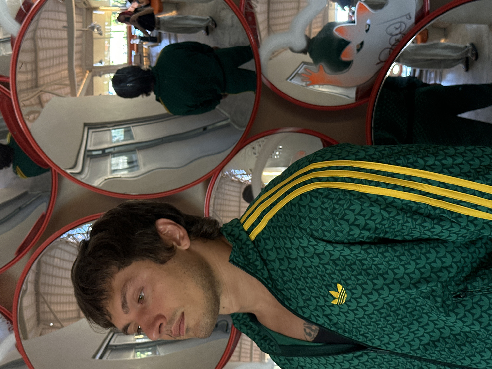 The image shows a man with light skin and dark brown hair, partially reflected in a round mirror. He is wearing a green Adidas track jacket with a leaf-like pattern and yellow stripes on the shoulders. A tattoo is visible on his upper chest. The mirror is part of a multi-mirror installation with red frames. Reflections in the other mirrors show parts of the surrounding environment, including a person in dark clothing and an inflatable cartoon figure. The location is identified as North Kuta, Indonesia based on the geotags.