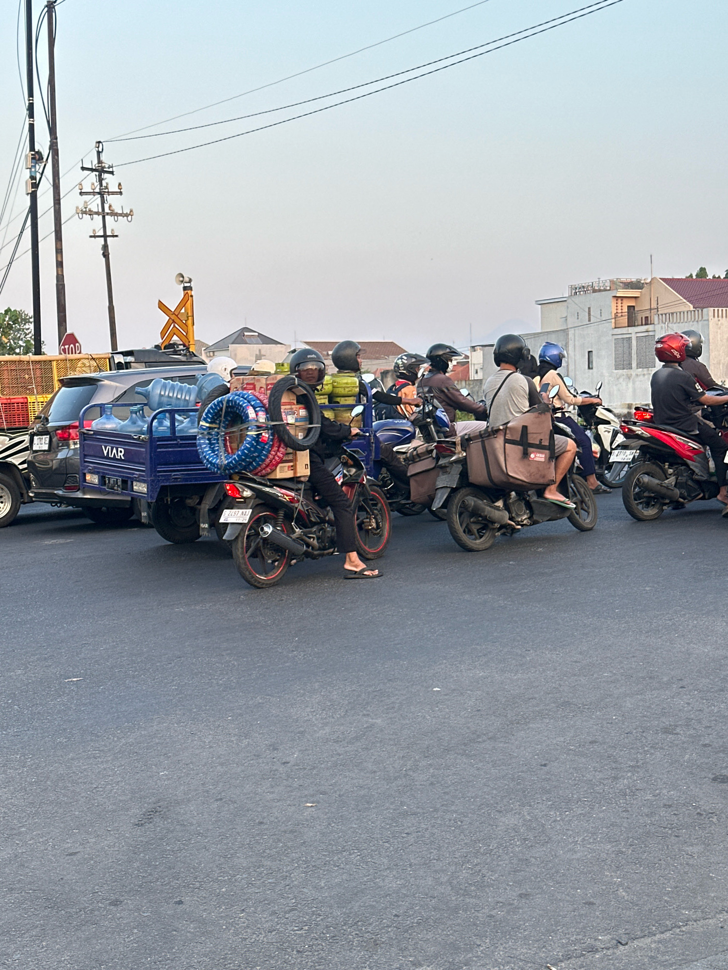 The image depicts a traffic scene in Mojokerto, Indonesia, characterized by a dense gathering of motorcycles and a small truck. The sky is clear and bright, suggesting daylight, possibly late afternoon given the warm lighting. The road surface is asphalt, and the surrounding environment includes residential buildings with tiled roofs and utility poles with overhead wires, typical of Indonesian urban or suburban settings.
In the foreground, several motorcycles are stopped or moving slowly. One motorcycle, black with red accents, is ridden by a person wearing a black helmet and dark clothing, with their feet bare, wearing flip-flops. This motorcycle is laden with cargo, including two tires mounted vertically and a larger, brown bag strapped behind the rider. Another motorcycle, also with a rider wearing a helmet, is positioned to the right of the first, with a red helmet visible. Further to the right, another motorcycle carries a rider with a black helmet and a passenger also wearing a helmet.
To the left, a small blue truck, branded "VIAR", is loaded with large water jugs. This truck is being followed by a dark-colored car, partially visible. A traffic signal with a "STOP" sign is visible on a pole to the left, indicating a controlled intersection. The overall impression is of a busy street with a significant volume of motorcycle traffic, common in Indonesian cities. The people are engaged in their daily commutes or delivery activities, with an emphasis on the practical use of motorcycles for transporting goods and people.