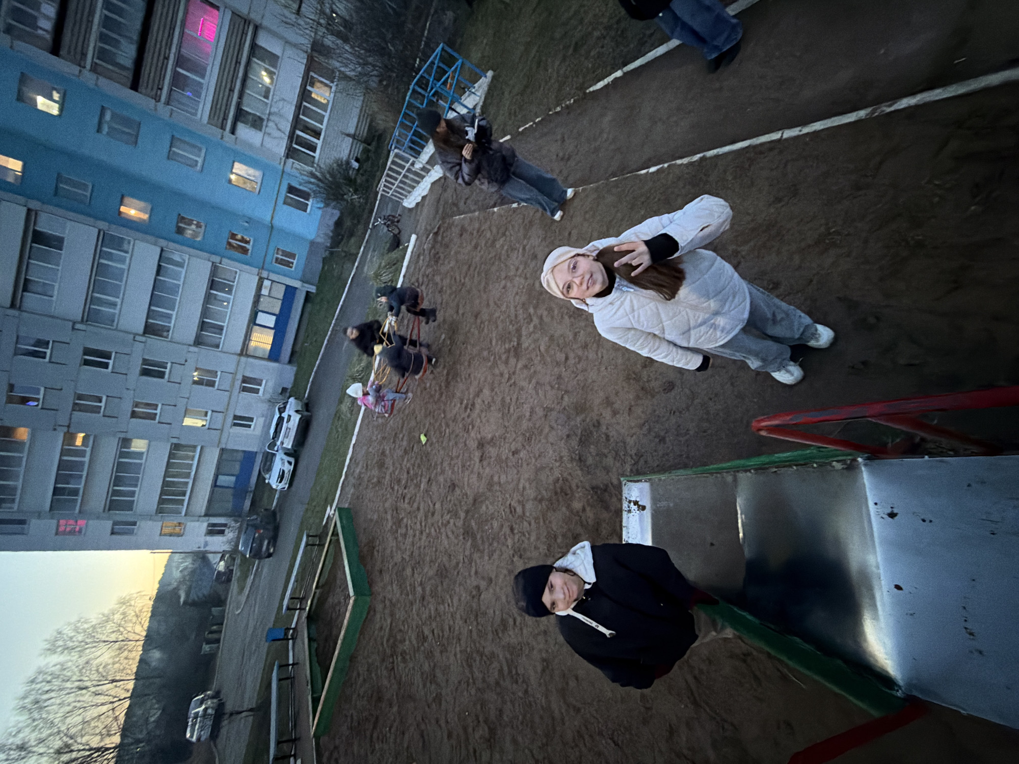 This high-angle image captures a group of young people gathered on a dirt playground during what appears to be dusk or early evening. The scene is set in an urban residential area, with a large, multi-story apartment building serving as a prominent background element on the left.
Several individuals are visible, all dressed in warm, casual attire, including puffer jackets, beanies, and pants, suggesting cool temperatures. In the mid-ground, a young woman in a white quilted jacket and a light-colored beanie smiles directly at the camera. To her right, in the foreground, another person in a dark jacket and black beanie stands beside a green-framed metal slide, also looking upwards. Further back on the left, other figures are seen interacting with playground equipment, including a blue metal climbing frame and smaller ride-on spring toys. A small yellow ball is visible on the ground in the mid-left.
The playground has a dark, earthy surface. The apartment building behind it features sections of light blue and grey facade with numerous rectangular windows. Many of these windows are illuminated with interior lights, some casting distinct colors like pink and orange, confirming the evening hour. Cars are parked along a road in front of the building. In the far background, bare trees stand against a muted sky, with a hint of natural light on the horizon. The overall impression is a casual, social gathering of friends in an everyday setting. No discernible text is visible in the image.