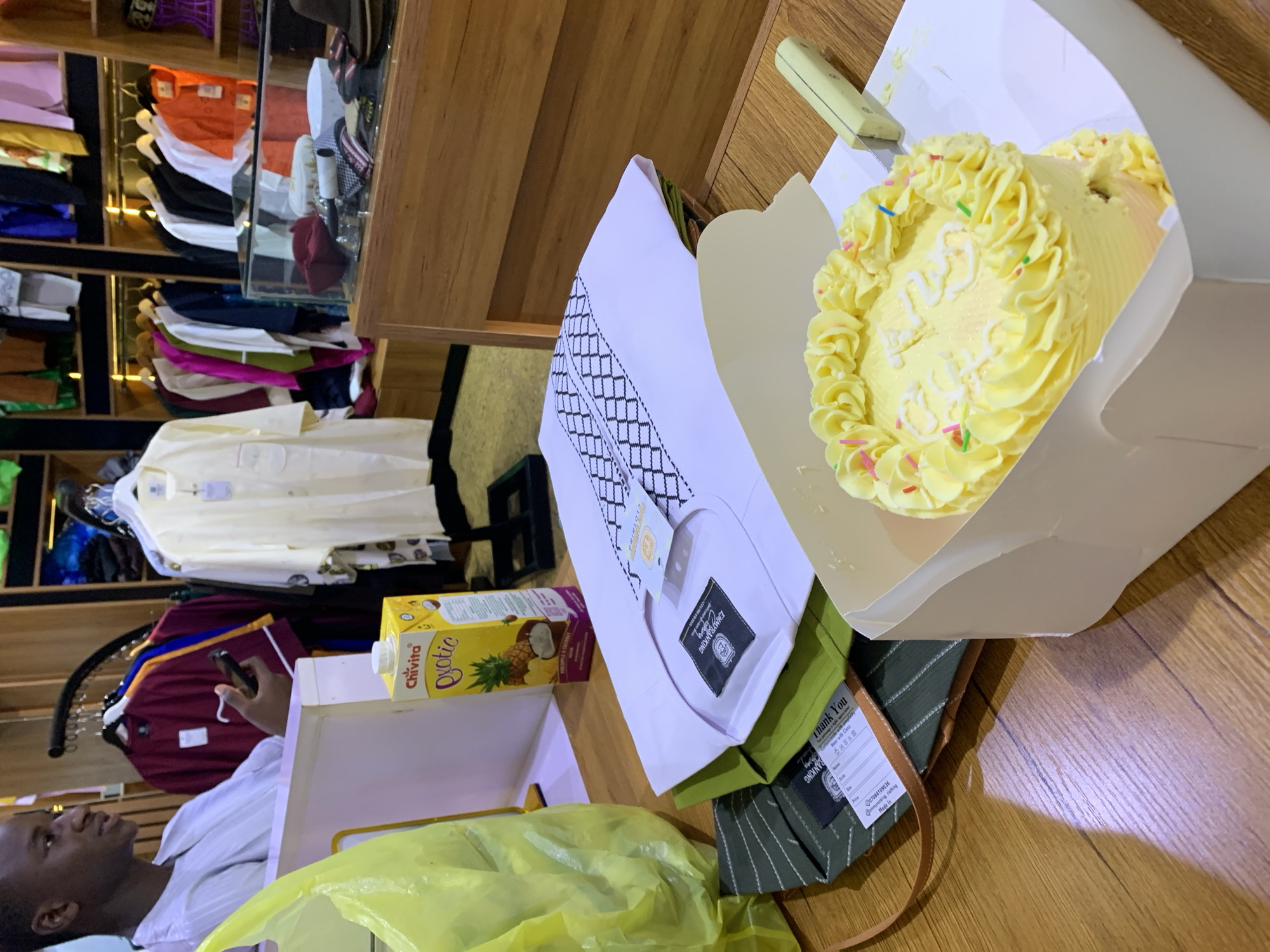 The image captures a close-up, eye-level view of a wooden table in what appears to be a clothing store in Gura Top, Nigeria. The foreground is dominated by a small, round, yellow cake with white frosting swirls and colorful sprinkles, placed in a white cardboard box. A light-colored knife with a yellow handle is resting on the right side of the box. Partially visible behind the cake are folded garments, including a white shirt with black embroidered patterns and a green striped garment. A yellow plastic bag is on the left, and a carton of Chivita Exotic juice with pineapple and coconut imagery is also on the table.
In the background, a young Black male, dressed in white clothing, is seated and looking to his left, holding a smartphone. Behind him, racks of clothing are displayed, featuring various colored garments, including burgundy, yellow, white, and orange shirts. Some garments are displayed on hangers, while others are folded on shelves. A glass display case with Islamic hats and other accessories is also visible. The overall lighting suggests an indoor environment, possibly a boutique or a retail establishment catering to traditional attire.