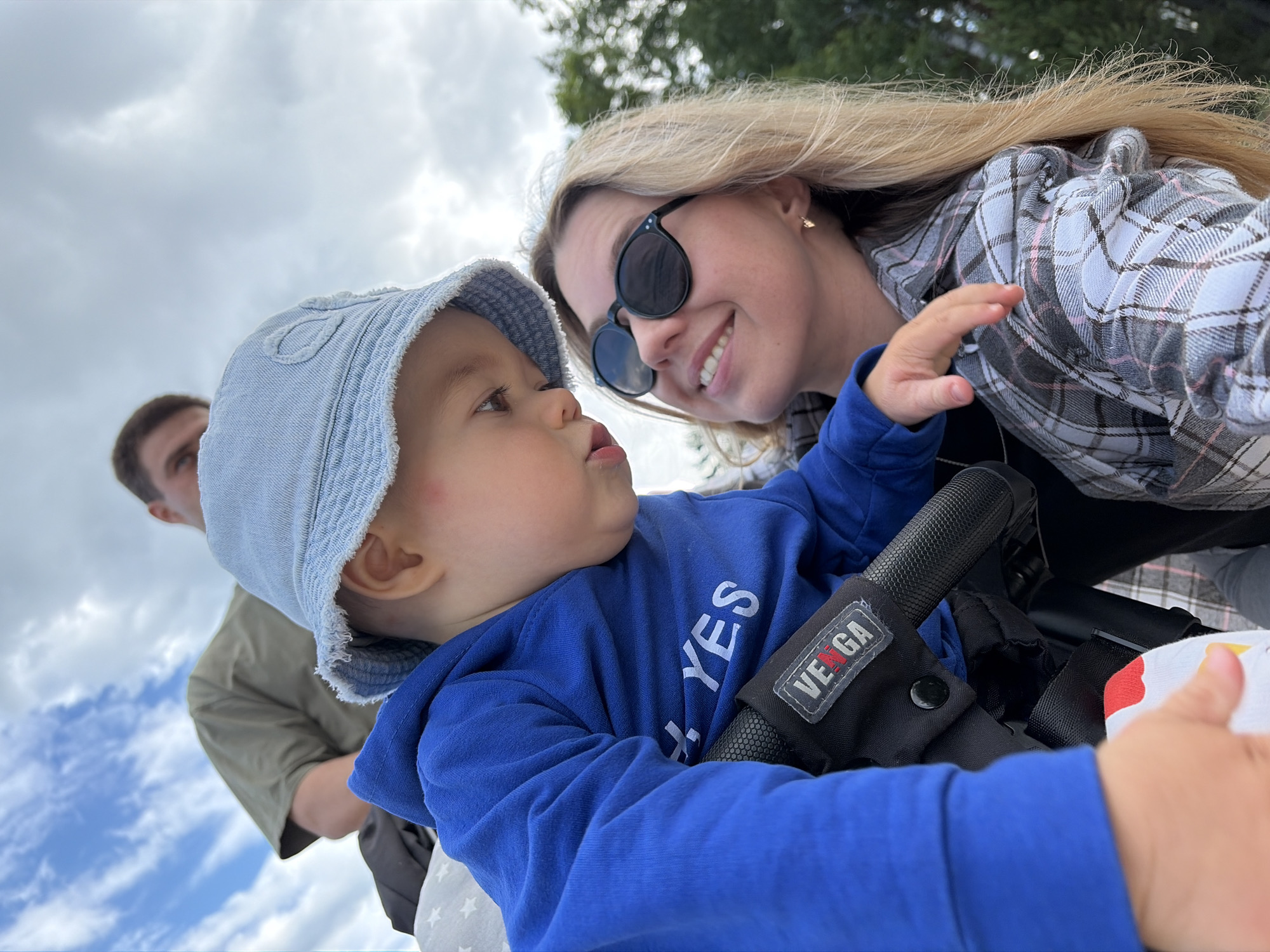 The image captures a tender moment between a mother and her young child, framed against a cloudy sky. The mother, with blonde hair and sunglasses, smiles warmly as she looks up at the child. The child, wearing a blue hoodie and a light blue bucket hat, is looking down and slightly to the side, with a red mark visible on its cheek. The child is being held in a stroller, with the brand "VEGA" visible on a tag. In the background, a man in a green t-shirt is partially visible, looking towards the camera. The environment appears to be outdoors, possibly a park or recreational area, given the presence of trees in the background. There are no specific landmarks or indicators within the image that would suggest the location is Chaikovsky, Russia.