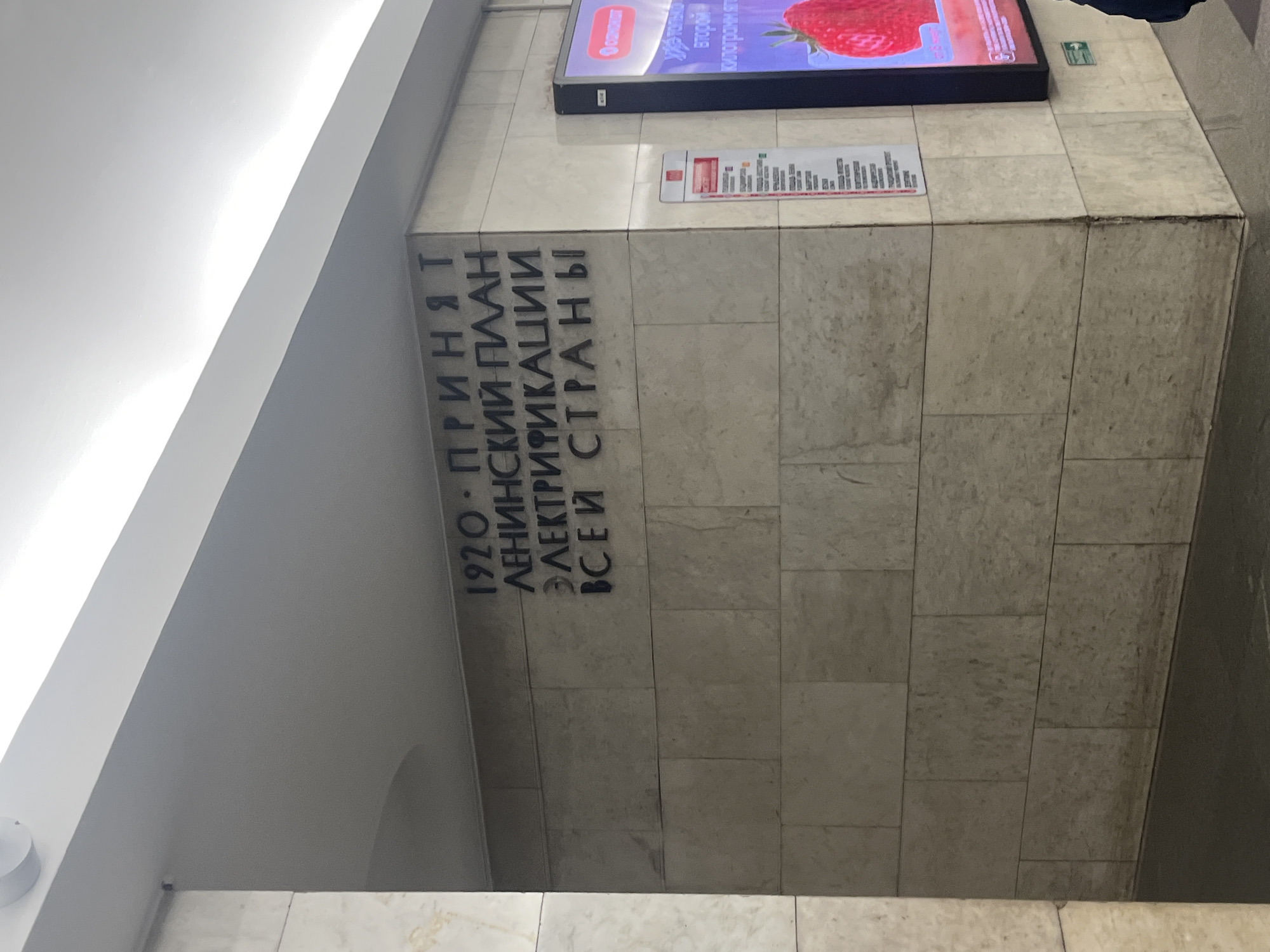 The image depicts a wall inside a metro station in Saint Petersburg, Russia. The wall is clad with light-colored marble tiles. Inscribed on the wall in dark lettering is the Russian text "1920 - ПРИНЯТ ЛЕНИНСКИЙ ПЛАН ЭЛЕКТРИФИКАЦИИ ВСЕЙ СТРАНЫ," which translates to "1920 - LENIN'S PLAN FOR THE ELECTRIFICATION OF THE ENTIRE COUNTRY WAS ADOPTED." Above the inscription, there is an advertising display screen with a partially visible image of a strawberry. Below this is a smaller display unit containing a map. The lighting is bright. The scene is indoors, likely a metro station due to the historical reference and typical metro station architecture.