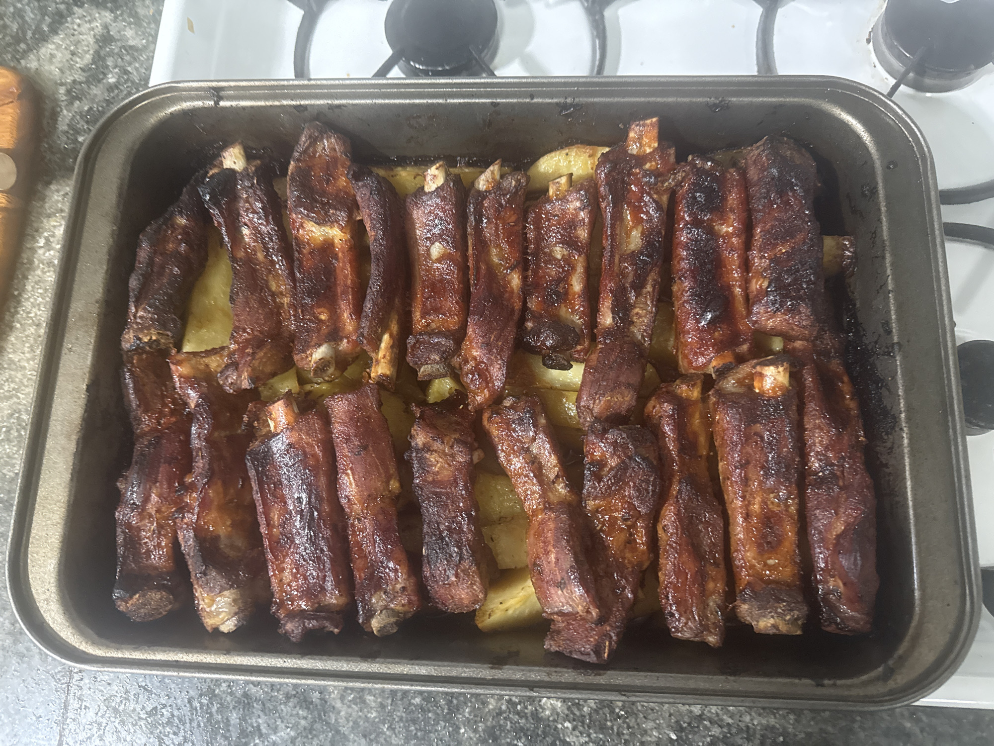 The image shows a rectangular baking tray filled with cooked ribs and potatoes. The ribs are arranged in rows, and they appear to be glazed or coated in a dark sauce. The potatoes are cut into chunks and are visible beneath the ribs. The tray sits on a stove top, with the burner knobs visible in the background. The scene is located in a kitchen.
