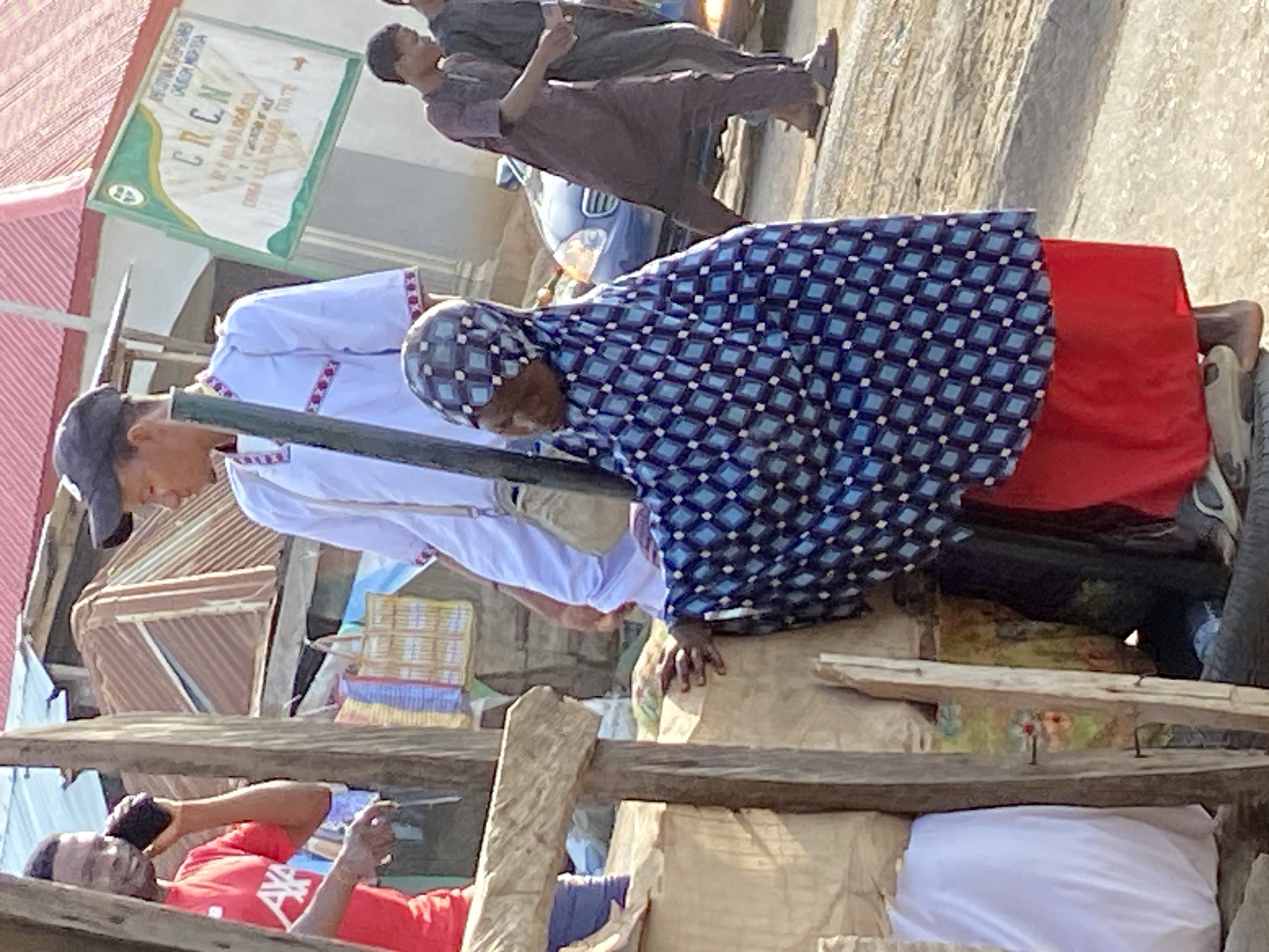 The image, rotated clockwise, captures a lively outdoor scene, likely a street or market in Mararaba, Nigeria, under bright daylight.
In the mid-ground, two individuals are prominent. To the left, a person wearing a white shirt with red and purple patterned trim, a dark cap, and light trousers, leans forward, looking down. Adjacent to them, another individual wears a blue and white diamond-patterned head covering and matching top, paired with a solid red skirt or wrapper. They are also leaning forward, with their right hand visible on a surface, suggesting interaction with a stall or goods. Behind these figures, several large, light-colored bags are stacked.
Below and slightly to the left, partially obscured by wooden planks, a person in a red t-shirt is visible, appearing to recline or lie down while holding a dark-colored phone to their ear.
In the background, two men are walking. The man closer to the foreground wears a dark, possibly long, traditional garment and appears to be holding a phone. Further back, another man in a dark shirt and trousers walks past a partially visible blue car.
A wooden framework, possibly part of a market stall or temporary structure, spans the foreground and mid-ground. On a building in the background, a green and white sign is mounted. The most prominent text on the sign is "CRCN". Above this, smaller, less legible text includes what appears to be "GENERAL CONFERENCE" and "CHURCH" or "MISSION". Below "CRCN", further text is visible but indistinct, possibly including "LADIES" and "YOUTH FELLOWSHIP" and a reference to "MARARABA".