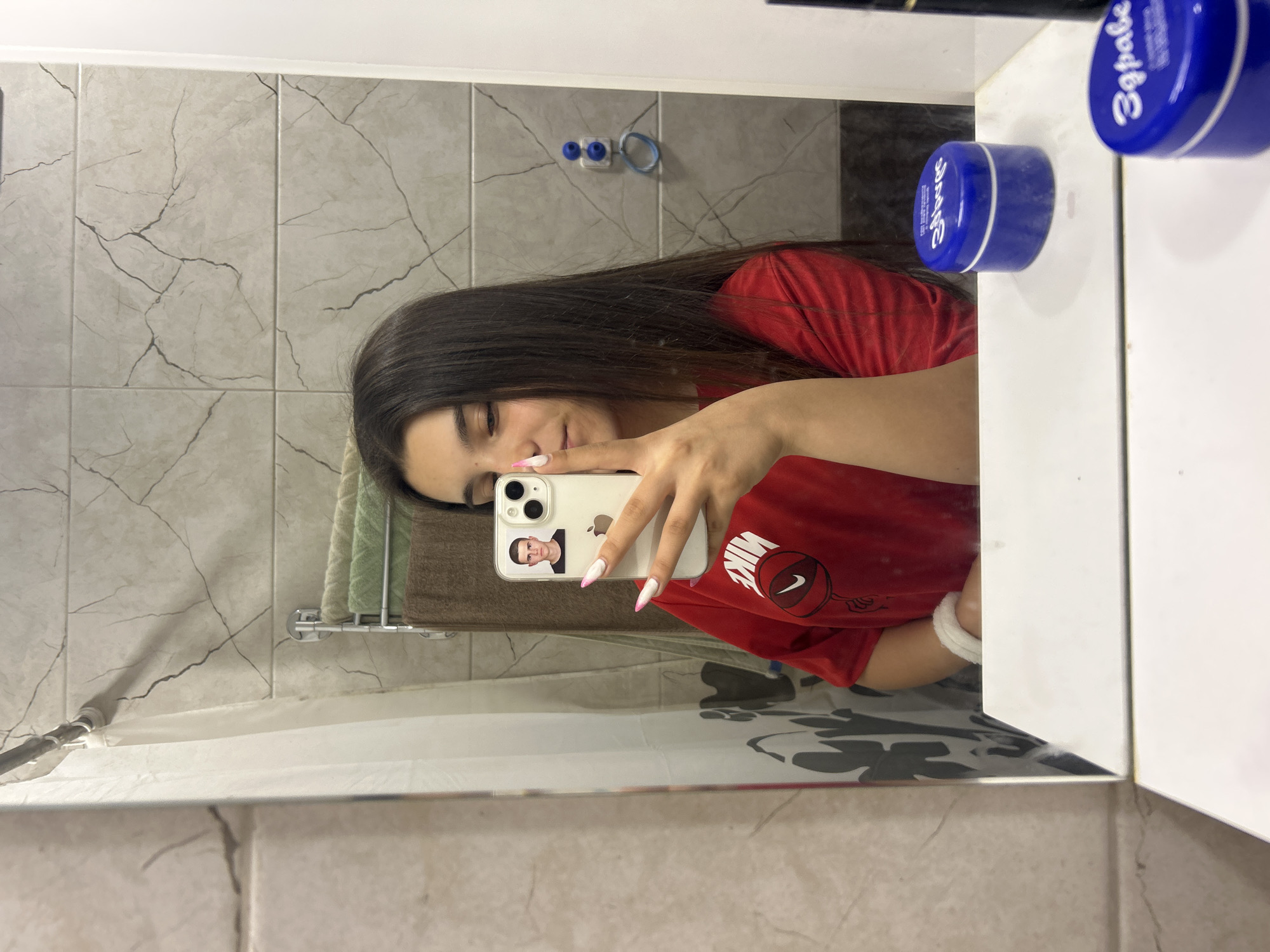 The image shows a young woman taking a mirror selfie in what appears to be a bathroom. She has long, dark hair and is wearing a red t-shirt. She is holding a white iPhone in her right hand, which is angled towards the mirror. The phone screen displays a selfie of a young man with short, dark hair. The woman's nails are painted with a pink and white design. On the counter in front of the mirror, there are two blue jars with white lids, one of which has text that appears to be in Cyrillic. The background shows a tiled wall with a shower curtain and a towel rack with towels. The scene is consistent with a bathroom environment. There is no information in the image to suggest the location is Shumen, Bulgaria.