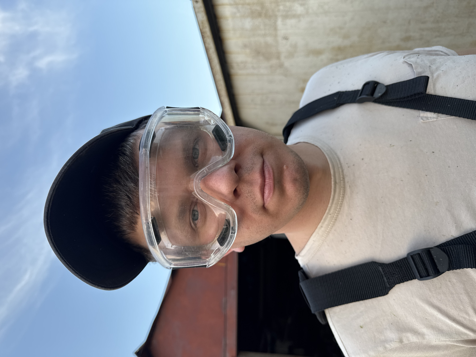 A man wearing clear safety goggles and a black baseball cap is looking directly at the camera. He is wearing a white t-shirt and black straps across his shoulder. The sky is visible in the background. The location is Ферма, Russia.