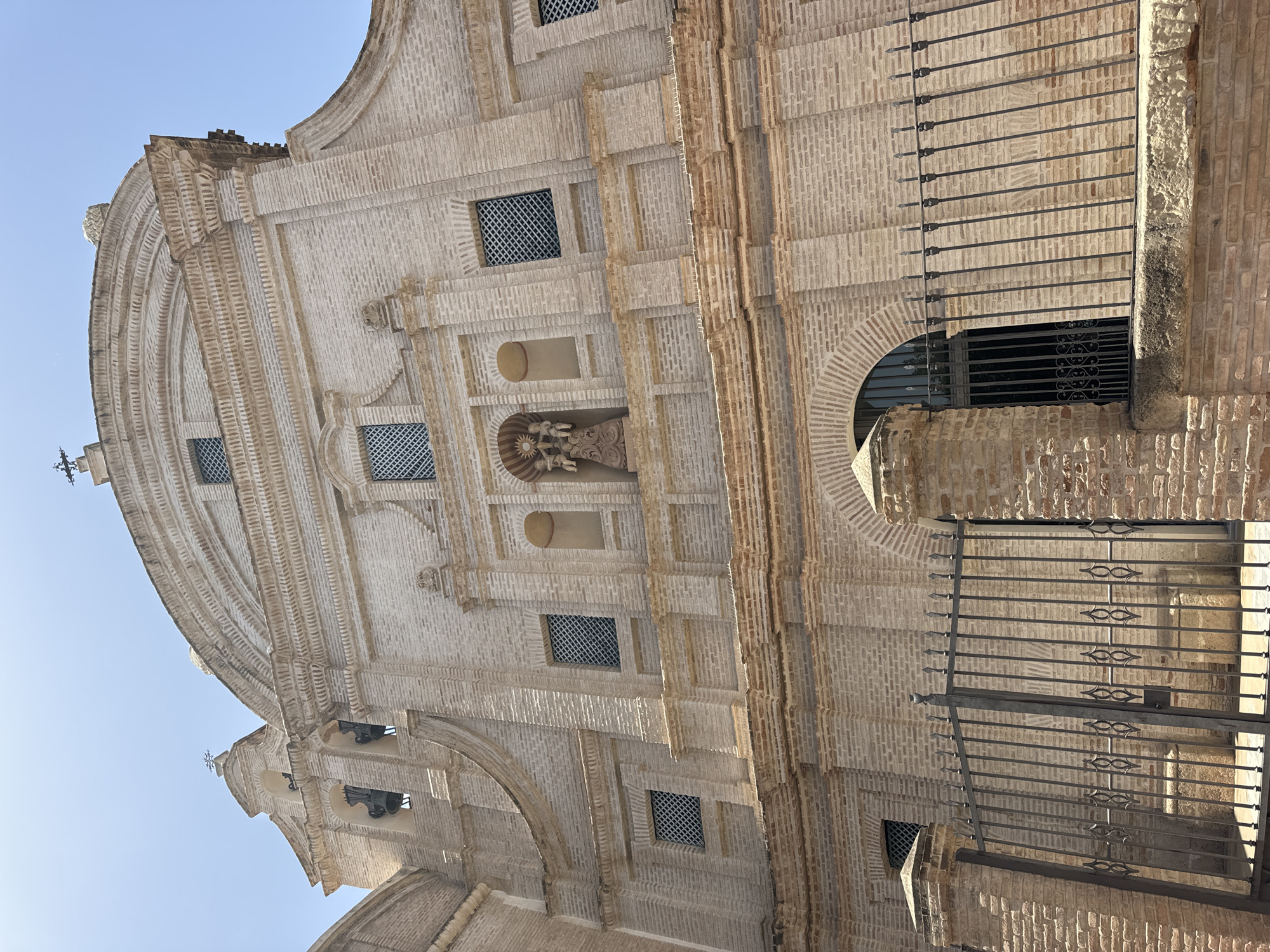 The image shows the facade of a historic brick building, likely a church, in Murcia, Spain. The architecture features arched windows with decorative grates, ornate brickwork with horizontal and vertical detailing, and a central niche containing a statue of religious figures. A wrought-iron fence with pointed finials runs along the bottom of the image, in front of a large arched entryway. The sky is clear and blue, suggesting a sunny day. The building's pale brick color stands out against the bright sky. The style appears to be Baroque or Renaissance, given the decorative elements and the overall grandeur.