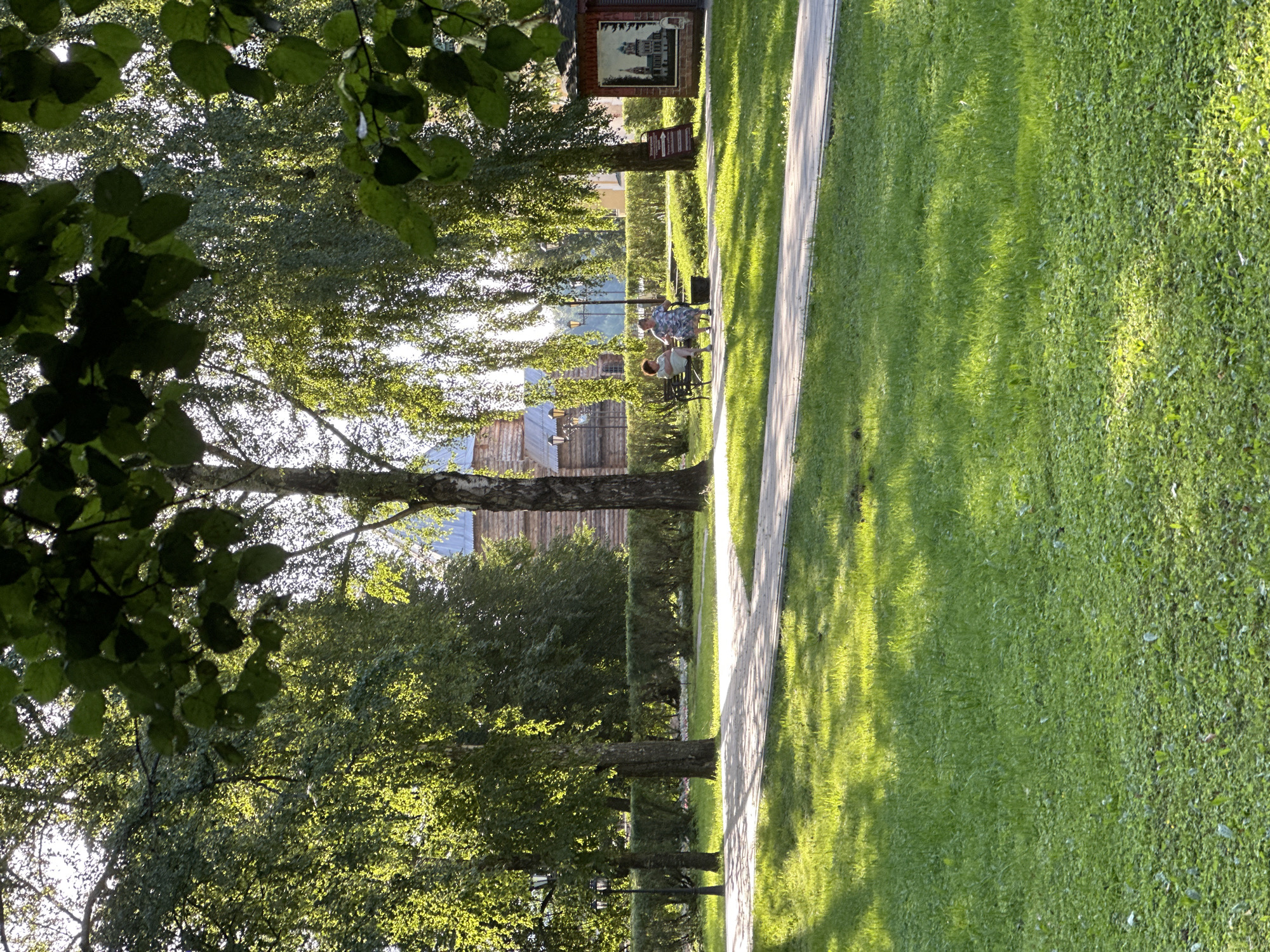 The image depicts a park-like setting in Selivanovo, Russia, characterized by lush green grass, paved pathways, and mature trees with dappled sunlight filtering through their leaves. In the background, a rustic wooden building, possibly a museum or historical structure, is visible. Two individuals, an older woman wearing a floral dress and another woman in a lighter-colored outfit, are seated on a park bench, appearing to engage in conversation. A small, red kiosk with a mural of a church on its side is positioned to the right of the bench. The scene evokes a peaceful and leisurely atmosphere, likely during daytime.