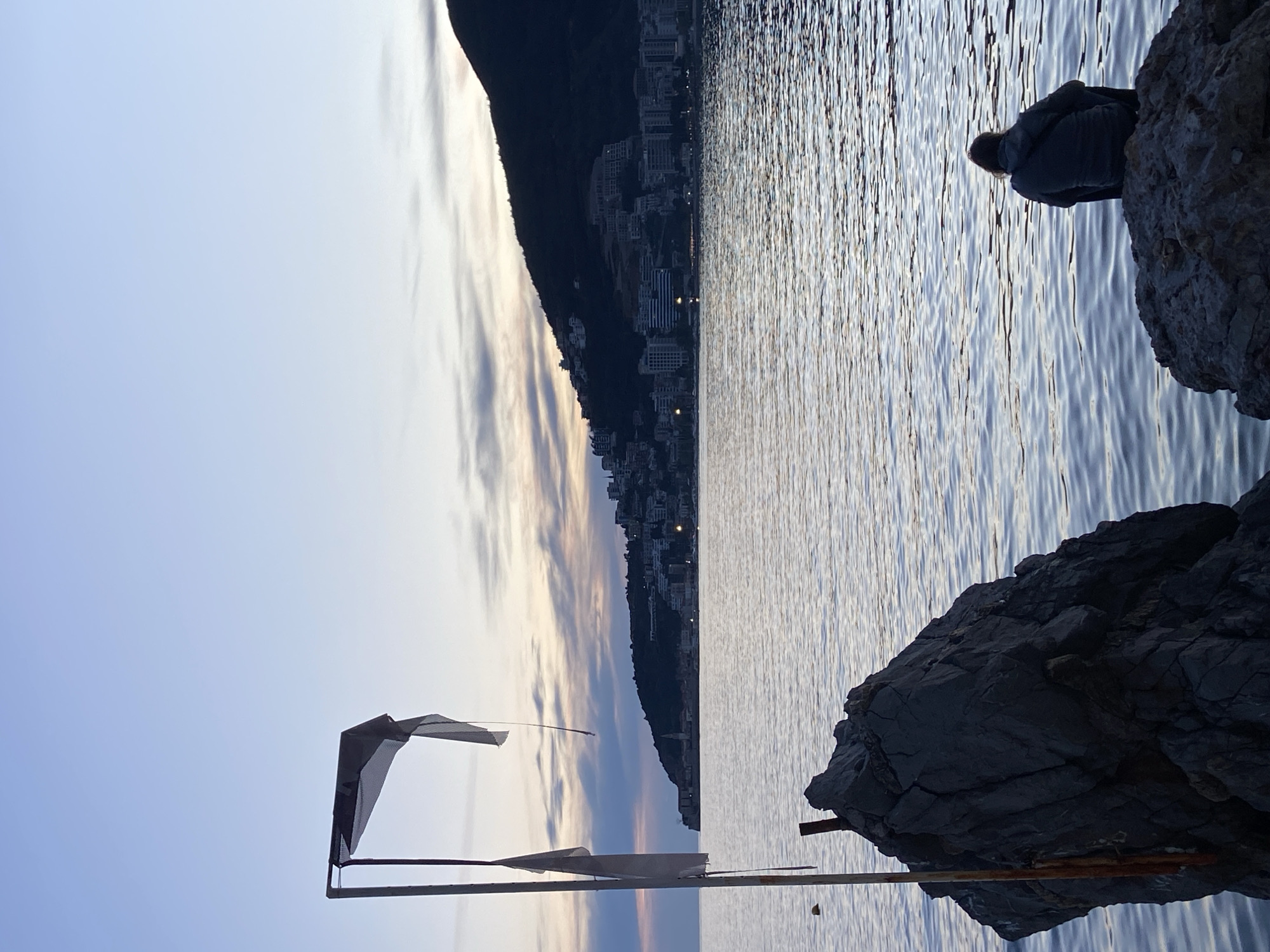 The image captures a coastline scene in Budva, Montenegro, at dusk or dawn. A person is sitting on a rocky outcrop overlooking the Adriatic Sea. The water has a slightly rippled surface, reflecting the sky. In the background is a mountainous landscape densely populated with buildings. In the foreground, a derelict structure stands, with elements like flags or fabric attached. The sky transitions from light blue to hues of orange and purple.