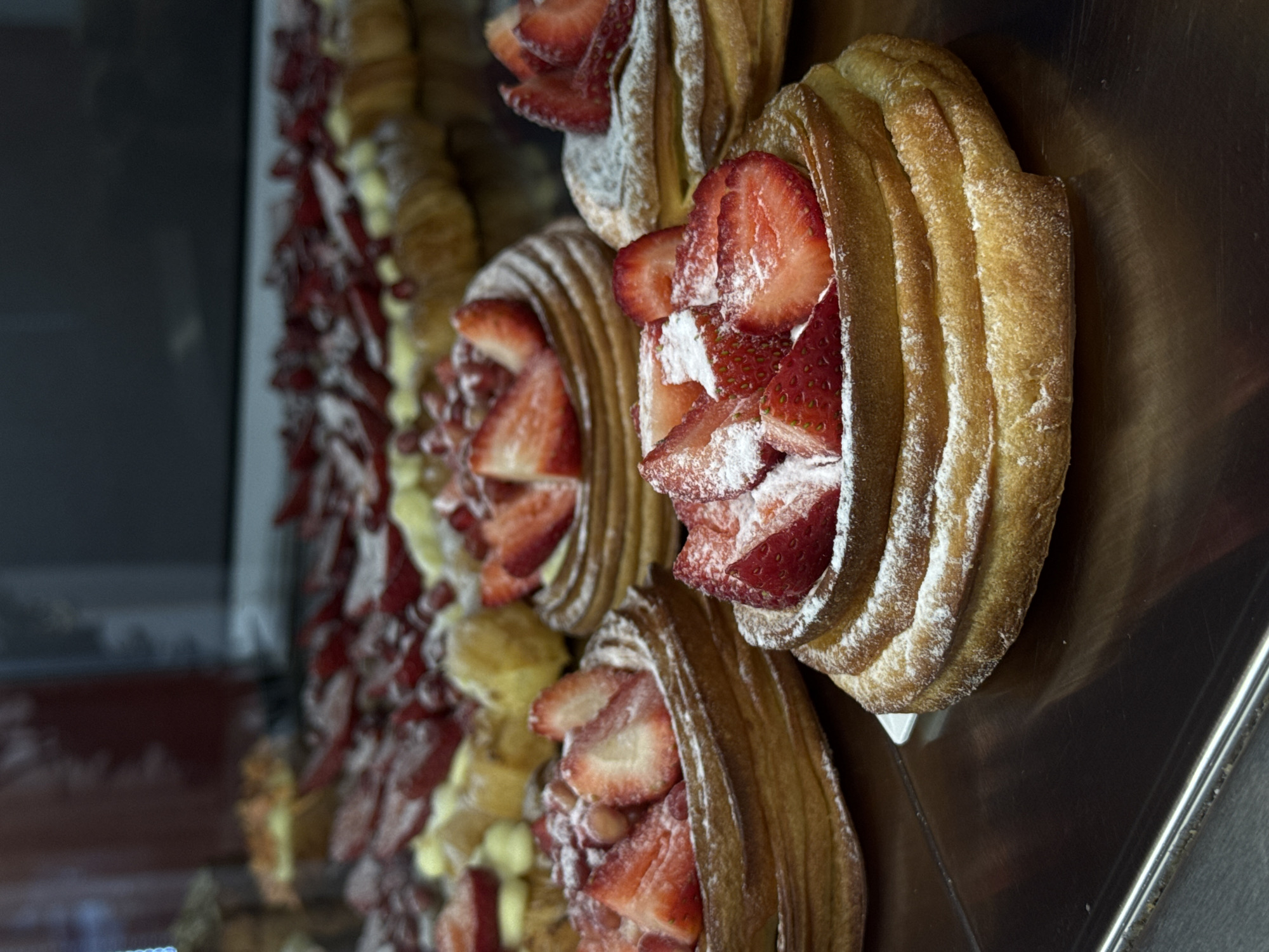 This close-up, vertically oriented image captures a display of appetizing pastries, likely in a bakery or patisserie in Yerevan, Armenia. The primary subjects are a row of golden-brown baked goods, possibly a type of choux pastry or cream puff, distinguished by their prominent ridged exterior. Each pastry is generously filled with numerous slices of vibrant, fresh red strawberries and delicately dusted with white powdered sugar, creating an appealing visual contrast.
The pastries are arranged on a dark, polished metallic surface that exhibits subtle reflections. In the softly blurred background, more similar pastries can be seen, extending in a line, suggesting a wider selection on display. The lighting appears to be artificial, effectively illuminating the textures of the pastries and the freshness of the fruit, enhancing their appeal. The scene depicts a typical commercial display of ready-to-sell sweet treats. There is no visible text within the image.