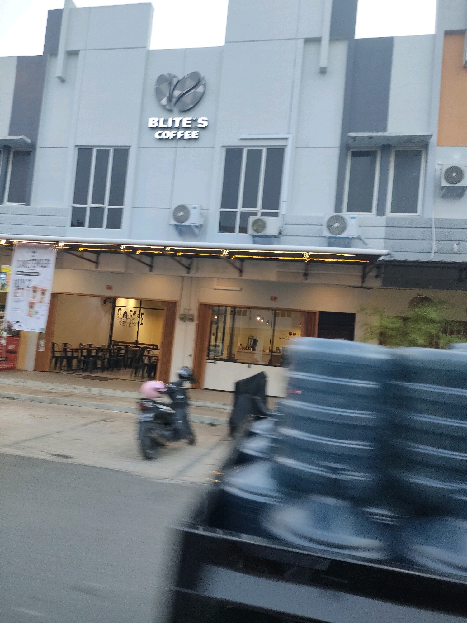 The image depicts the exterior of "BLITE'S COFFEE" in Batam, Indonesia. The establishment is a two-story building with a modern facade. The upper floor features several large, rectangular windows with dark frames and a sign for "BLITE'S COFFEE" with a coffee bean logo above it. Air conditioning units are visible mounted on the exterior wall. The ground floor has a covered walkway with illuminated signage and an entrance to the coffee shop, which reveals tables and chairs inside. A banner advertising "BUBBLE TEA" is hanging on the left side of the entrance. In the foreground, a motorcycle with a pink helmet is parked on the side of the street, and a portion of a vehicle carrying large blue water jugs is in the lower right corner, suggesting motion blur. The environment appears to be an urban street with a sidewalk.