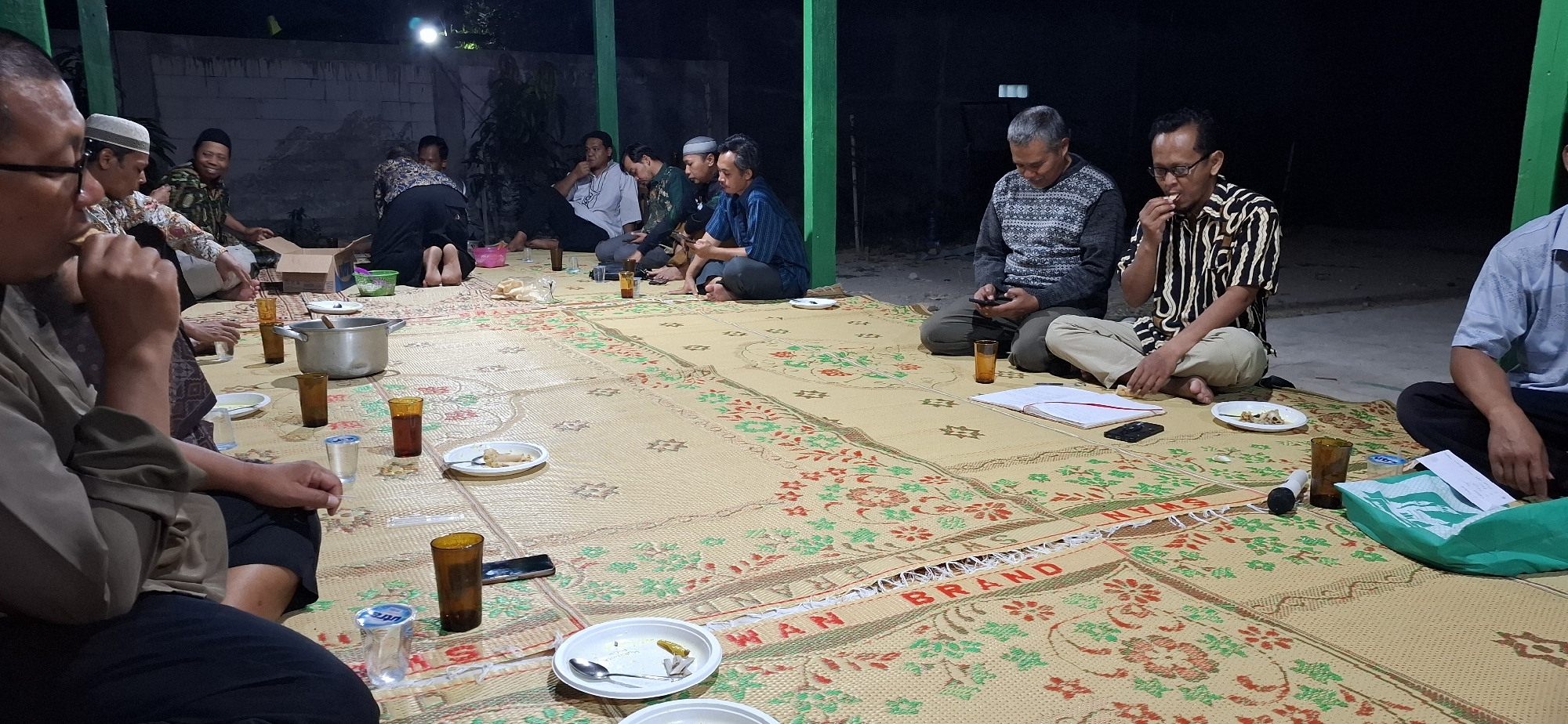This is a forensic, matter-of-fact description of a gathering in Wonorejo, Indonesia.
The scene depicts a group of at least nine adult males sitting on woven mats laid out on the ground, likely outdoors given the presence of a concrete wall and pillars. The gathering appears to be a meal or social event, possibly in the evening or night, as indicated by the dim lighting.
Several individuals are actively eating, with one man in the foreground to the left biting into food. Others are engaged in conversation or looking at their phones. The men are dressed in casual clothing, including shirts, trousers, and some are wearing skullcaps, suggesting a cultural or religious context.
Various objects are present on the mats, including plates with food remnants, glasses containing beverages, a large metal pot, a cardboard box, a notebook, and what appears to be a microphone. A green plastic bag is also visible, suggesting take-out food or supplies. The woven mats themselves have a repeating pattern of floral designs in green and red on a beige background, with visible text branding "SWAN BRAND" on some sections.
The environment is simple, with a rough concrete wall in the background and visible pillars, one painted green. The overall impression is of an informal community gathering.