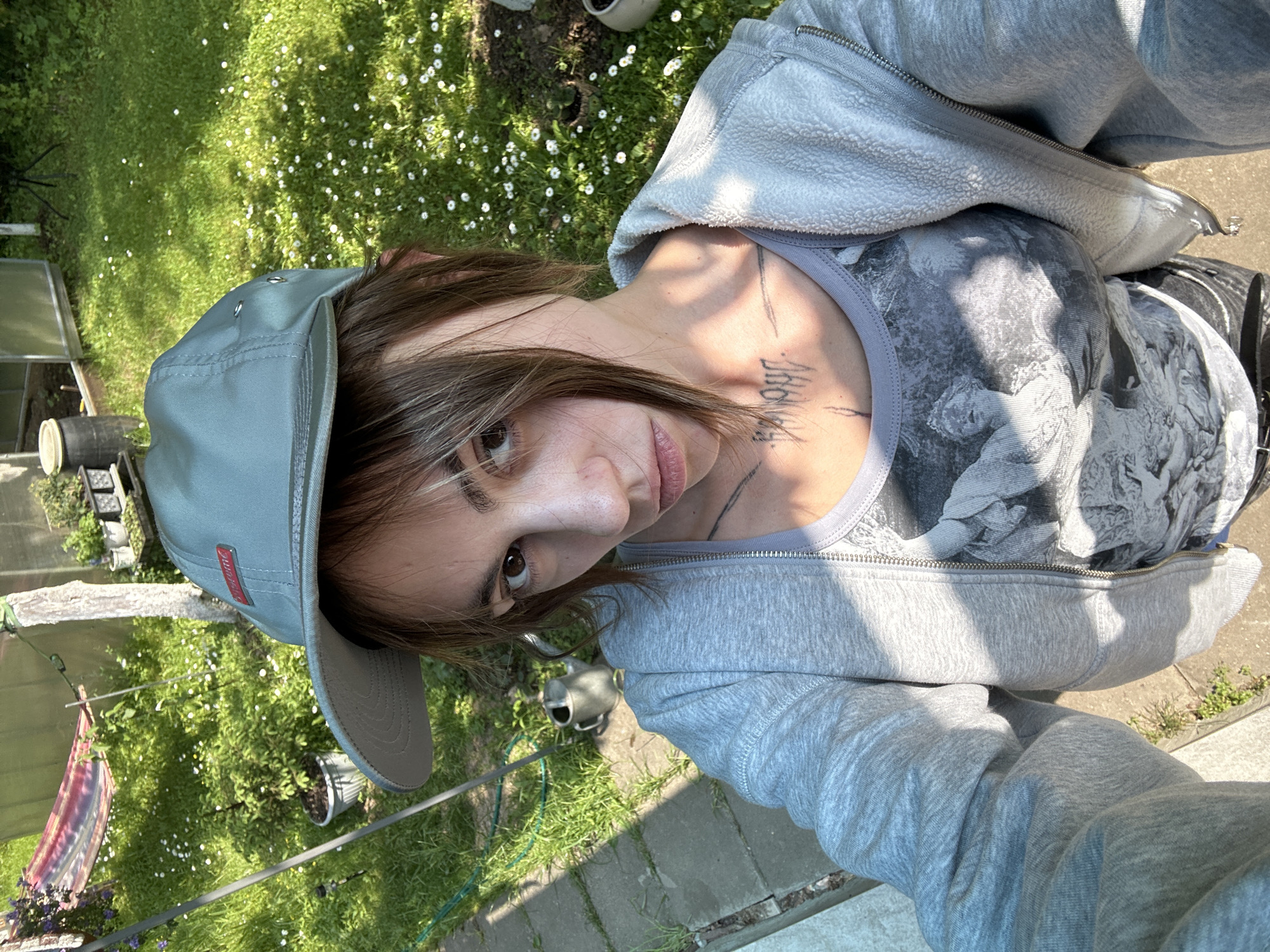 The image shows a young woman with brown hair, wearing a grey zip-up hoodie over a grey tank top with a black and white graphic print. She also has a light grey baseball cap on her head, with a red tag on the front. The woman has tattoos visible on her neck and chest. She is outdoors, in what appears to be a backyard or garden area. The background includes green grass with small white flowers, bushes, a hammock strung between two poles, a watering can, paving stones, and a greenhouse-like structure. The lighting suggests it is a sunny day. There is no visible indication of the location being in Козино, Russia.