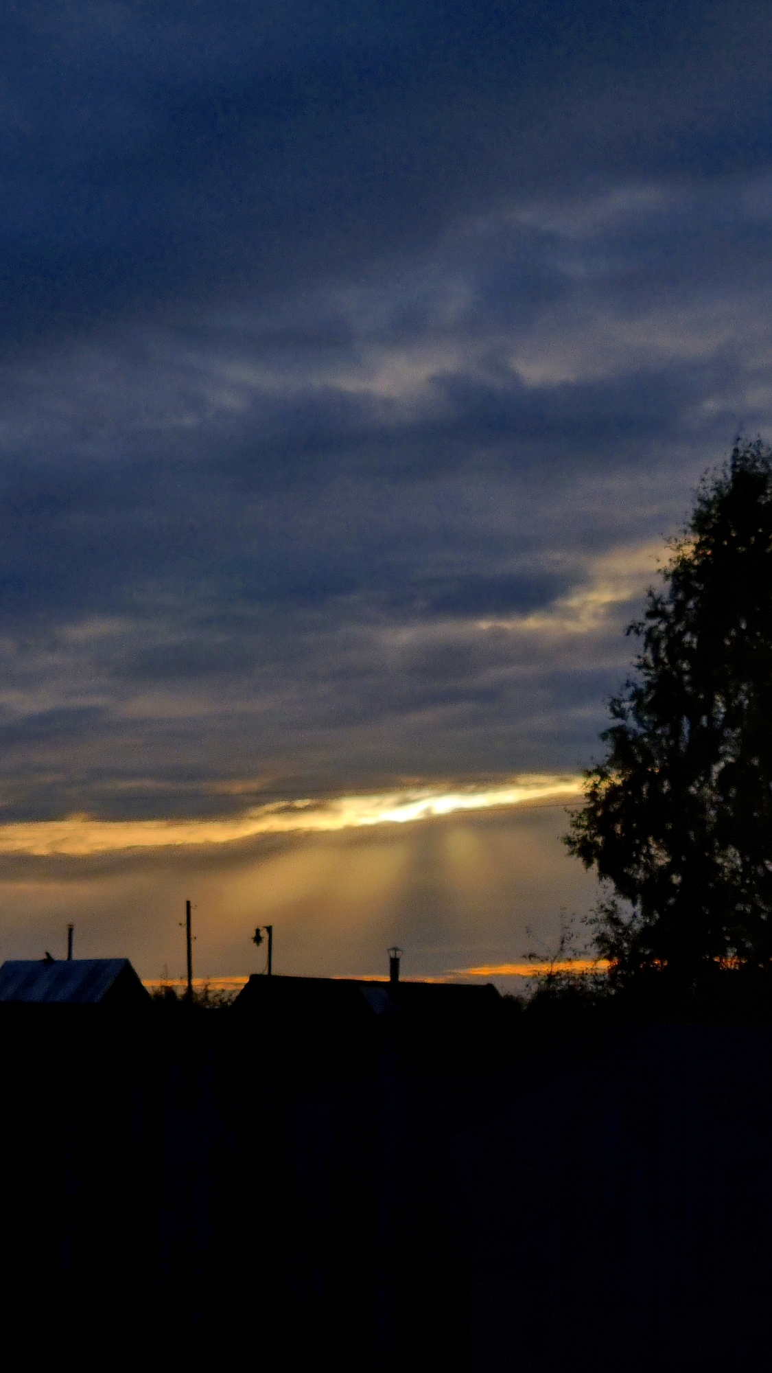 The image depicts a dark, cloudy sky over the city of Vysokoe, Russia, at dusk. The sky is a deep blue, with streaks of gray and lighter clouds. A bright band of orange-yellow light pierces through the clouds near the horizon, casting rays of light downwards. In the foreground, the dark silhouettes of several buildings and a tree are visible. One building on the left appears to be a small house with a slanted roof. To its right, a utility pole stands tall, followed by another building with a chimney. A large tree silhouette dominates the right side of the frame. The overall scene is characteristic of an evening in a rural or semi-rural area, with muted colors and a dramatic sky. There are no people or animals visible in the image.