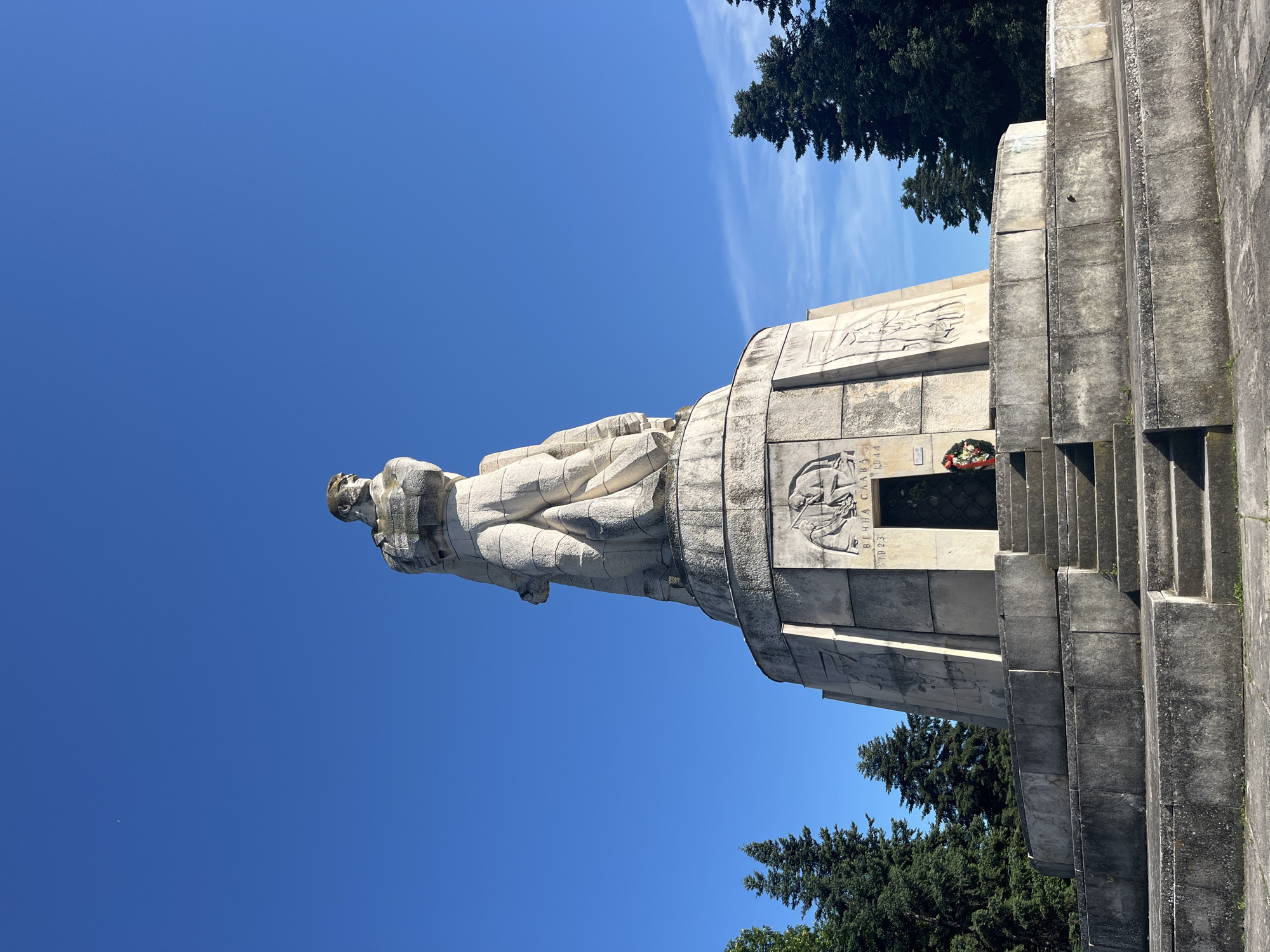 The image shows a monumental statue of a soldier standing atop a large, tiered base. The statue is made of stone and depicts a male figure in military attire, looking off to the side. The base of the monument is also stone and features reliefs and inscriptions, including the years "1923" and "1944" and the text "ВЕЧНА СЛАВА" (Eternal Glory). There is an open doorway at the base of the monument, with a wreath of red flowers hanging to the left of the entrance. A set of stone stairs leads up to the doorway. The monument is situated in an outdoor environment with a clear blue sky and several tall evergreen trees surrounding it. The overall impression is one of solemnity and remembrance. This monument is located in the city of Varna, Bulgaria and is known as the Pantheon of National Revival Heroes.