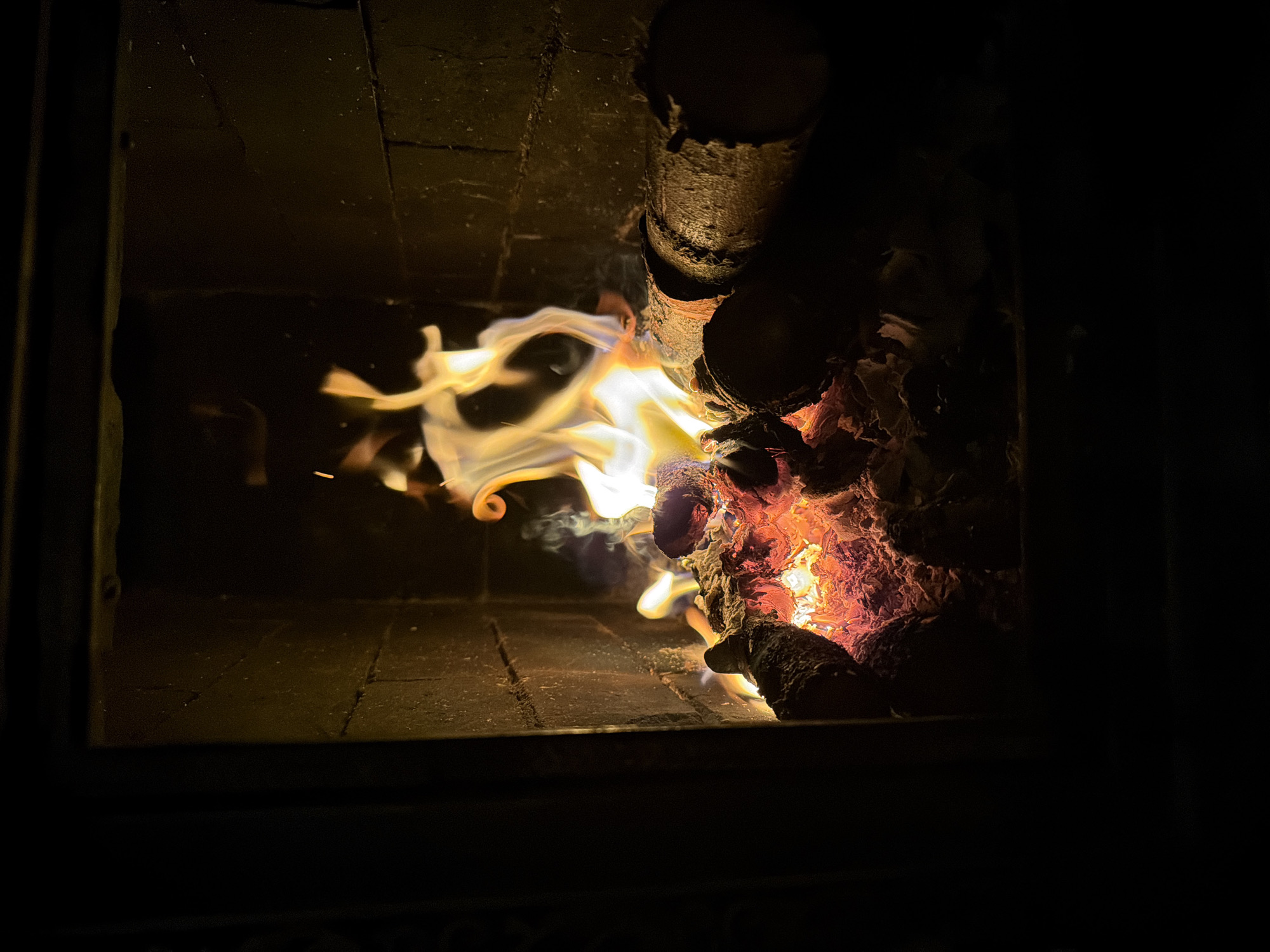 The image shows a fire burning inside a brick-lined fireplace or wood stove. The flames are yellowish-white and swirl around partially burnt logs. The wood on the right side is glowing with orange and red heat. The scene is dark, with the fire providing the main source of illumination. The bricks forming the interior of the stove are visible, showing signs of soot and wear. The location in the city of Пролетарский, Russia is not discernable from the content of the image.