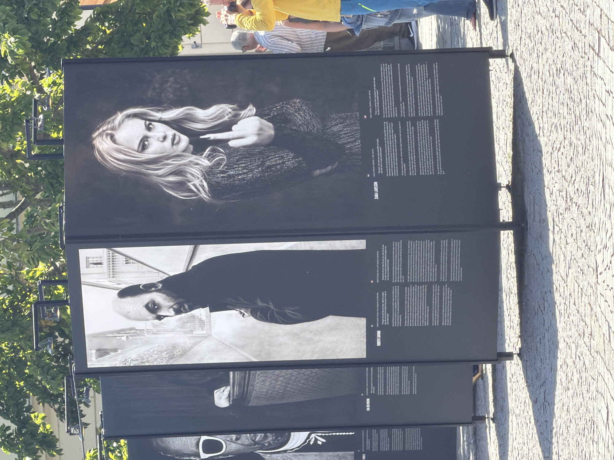 The image depicts an outdoor exhibition in Lund, Sweden, likely near Lund Cathedral due to the architectural style of the background buildings. The exhibition consists of several large black display panels, each featuring a black and white photograph and accompanying text.
The leftmost visible panel displays a portrait of a man wearing glasses, his face partially obscured. Next to it, a panel shows a man with a bald head, looking to the side, with text below. The third panel, prominently featured, shows a close-up of a woman, identified by text as Zara Larsson, giving the middle finger. Her expression is defiant. The fourth panel is mostly obscured but appears to be another portrait.
In the background, people are visible looking at the exhibition. One man in a yellow shirt is holding a camera to his face, as if photographing the art. Others are standing, observing the displays. The ground is paved with cobblestones, and the scene is bathed in sunlight, casting shadows from the display panels. The foliage of trees can be seen above the exhibition.