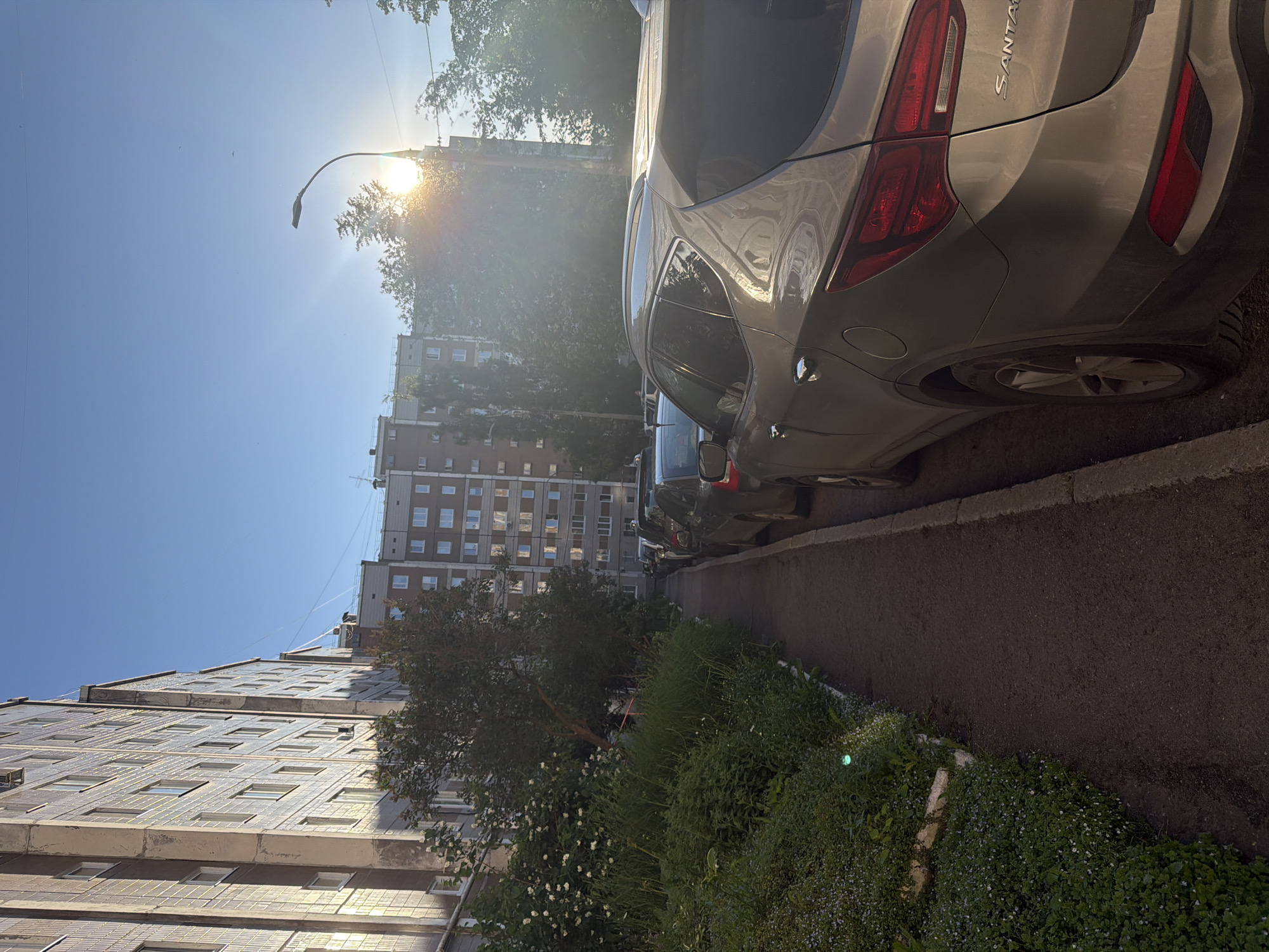 This is a vertical, low-angle outdoor shot of an urban scene on a sunny day in Izhevsk, Russia. The image is dominated by a light-colored SUV parked on the right side of the frame, facing away from the viewer. The SUV's rear is visible, with its taillights and "SANTA" badging clearly discernible. In the background, a tall, multi-story apartment building with a grid of windows stretches across the frame. To the left of the building, another similar building is partially visible. A street lamp with a curved arm arcs over the scene, with the bright sun positioned behind it, creating lens flare and casting rays of light through the leaves of trees. The sky is a clear, vibrant blue. A strip of vegetation, including bushes and small white flowers, runs along the edge of the asphalt road where the car is parked. The overall mood is bright and summery, suggesting a peaceful, everyday moment in a residential urban environment.