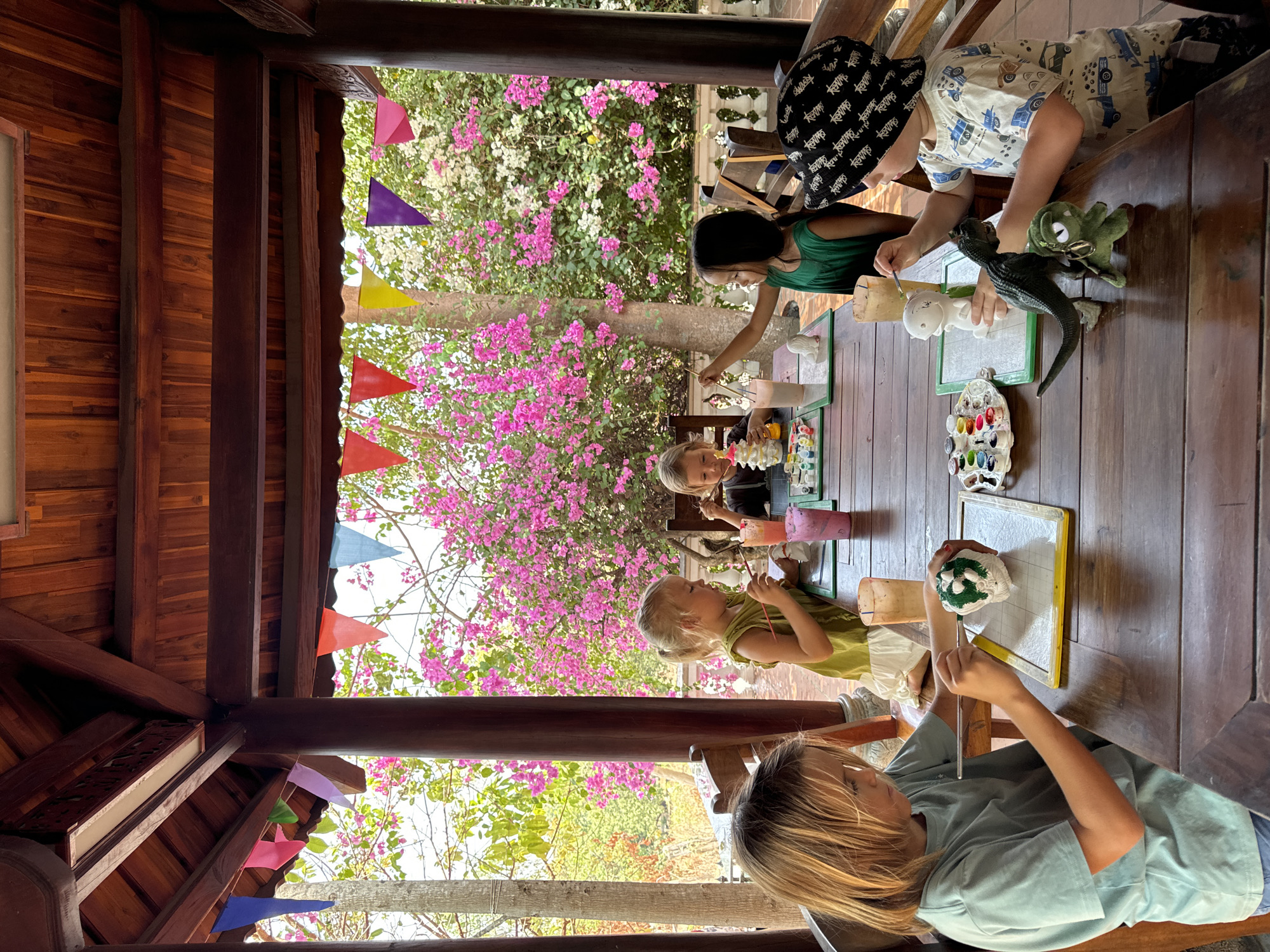 Five children are seated around a long wooden table, engaged in a painting activity. The setting appears to be an outdoor covered area, possibly a patio or veranda, as indicated by the wooden structure of the roof and posts. Brightly colored triangular flags are strung up, adding a festive touch to the scene.
The children are focused on painting small, unidentifiable objects placed on small whiteboards in front of them. Paint palettes and cups are scattered across the table. One child has a toy dinosaur next to his whiteboard.
The background features a profusion of blooming bougainvillea in shades of pink and white, with greenery visible beyond. The bright sunlight indicates a daytime scene.
The location is Mũi Né, Vietnam.