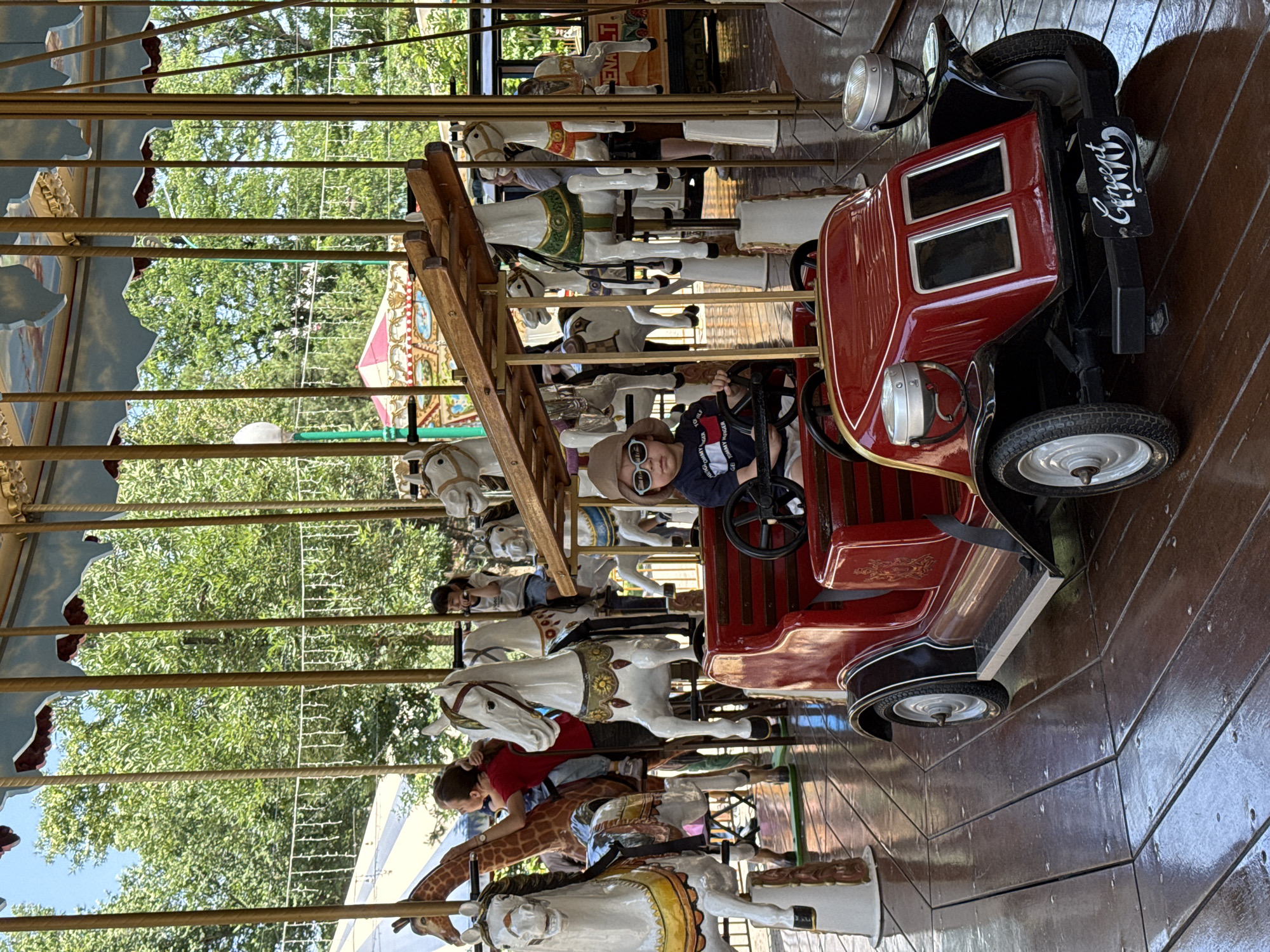 On a merry-go-round, a young child wearing a tan hat and sunglasses sits in a red vintage-style car. The merry-go-round features painted horses and a giraffe figure. Passengers are visible on some of the horses. The scene is outdoors, with trees and a structure visible in the background. The merry-go-round has a decorated canopy. The location is possibly in Tashkent, Uzbekistan.