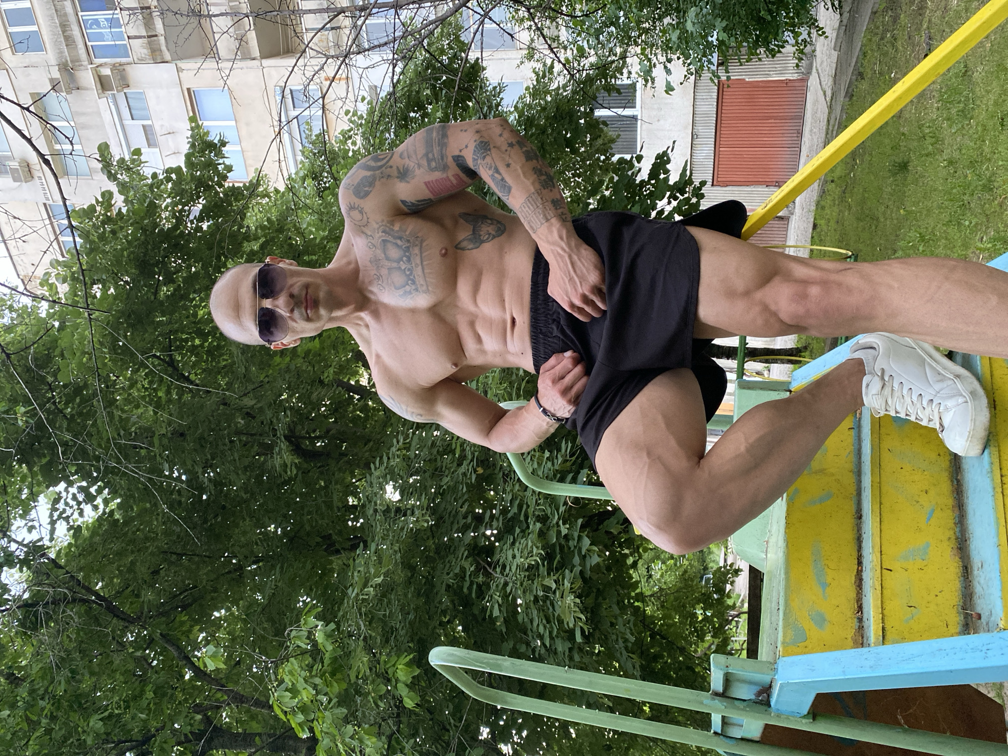 The photograph depicts a muscular, bald man wearing sunglasses, black shorts, and white sneakers. He is positioned on a yellow and blue playground structure, likely located in the city of Pleven, Bulgaria, judging by the background architecture. The man has numerous tattoos on his arms and torso. The background includes trees and a multi-story residential building. The image has been rotated, resulting in a tilted perspective.
