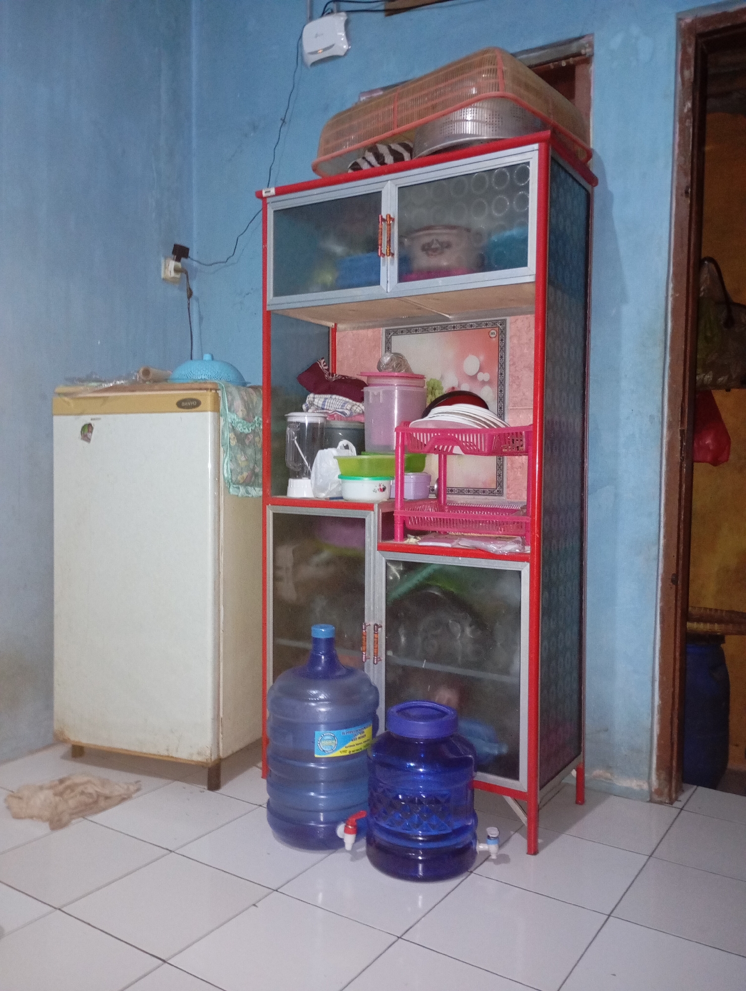 This is a medium shot of a living area in Kalitinggar, Indonesia. The walls are painted a light blue. In the center of the image is a tall, narrow, red-framed cabinet with glass doors. The cabinet contains various items, including plates, containers, and what appear to be food storage bags on the shelves. On top of the cabinet is a basket-like object, possibly for storing food or pet items. To the left of the cabinet is a white refrigerator. In front of the cabinet, on the tiled floor, are two large blue water dispensers with attached taps. One is slightly larger than the other and is positioned behind it. A small pile of light-colored material, possibly a rag or cloth, is on the floor to the left of the refrigerator. The overall scene depicts a functional living space with storage and utilities.