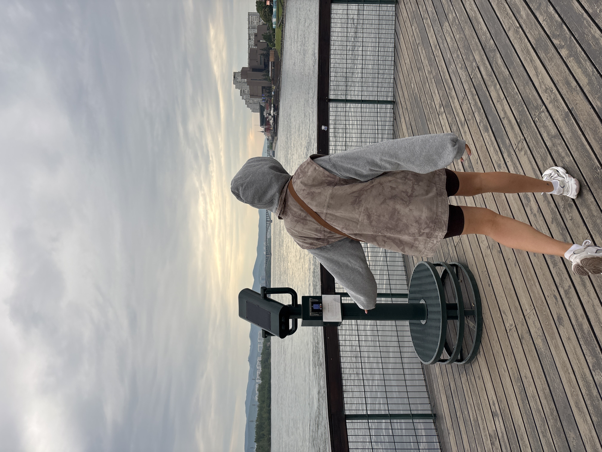 The image depicts a person from behind, standing on a wooden promenade overlooking a wide river. The person is wearing a grey hooded sweatshirt, a mottled brown vest, dark shorts, and white sneakers. In front of the person is a dark green viewing scope on a pedestal, with some informational signage attached. The river is calm, and in the distance, across the water, is a city skyline with numerous buildings and a bridge. The sky is overcast with soft, diffused light. The promenade is made of weathered wooden planks and is bordered by a metal railing. The location is the embankment of the Yenisei River in Krasnoyarsk, Russia, specifically the area near the viewing scope that offers panoramic views of the city and the river.