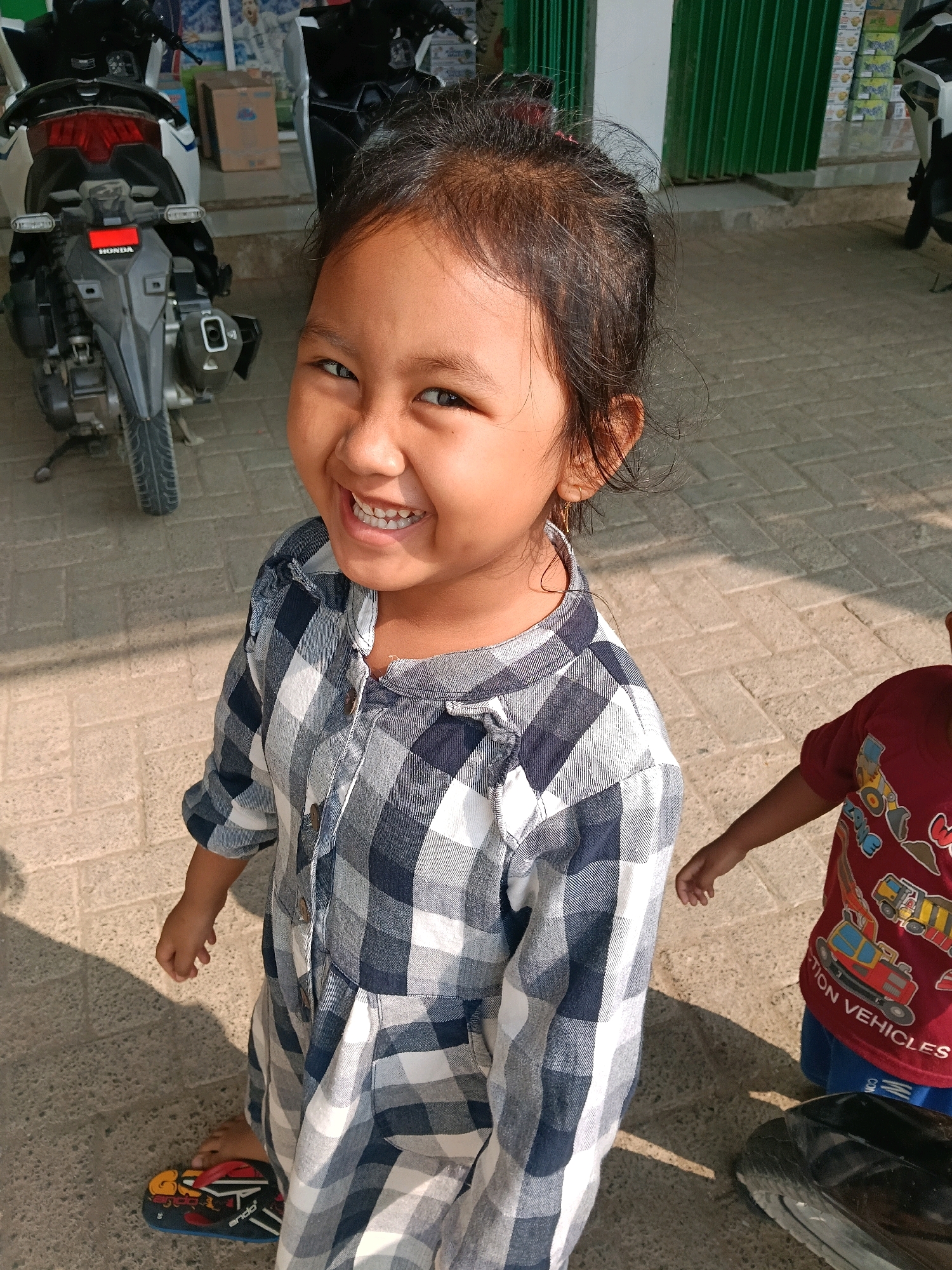 A young girl with dark hair and a wide smile stands outdoors, wearing a blue and white checkered shirt. She is looking at the camera. Her right ear has a thin, gold hoop earring. To her right, a portion of a second child, wearing a red shirt with "Construction Vehicles" printed on it, is visible. They are standing on a paved area with a shadow cast on it. In the background, a motorcycle is parked to the left, and a green shuttered building is behind the children. The scene appears to be in a city environment.
