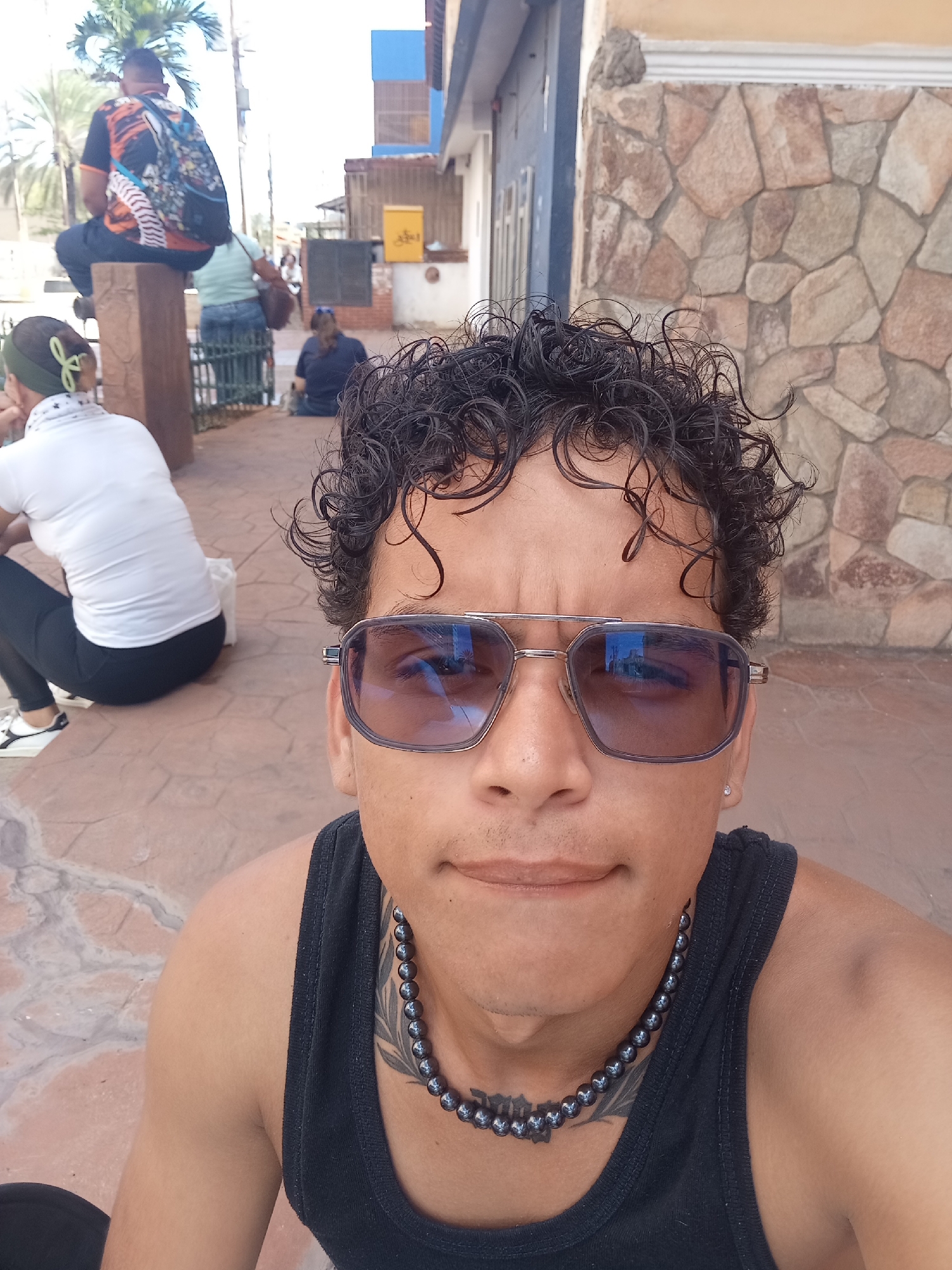In Juan Griego, Venezuela, a close-up selfie captures a person wearing sunglasses and a black tank top. The individual has curly dark hair and is wearing a beaded necklace. Behind them, other individuals are visible sitting outdoors. One person in the background is wearing a colorful t-shirt and backpack, seated on a wooden post. Another person in a white shirt and black leggings is sitting on the ground. The environment appears to be an outdoor public space with a stone-patterned wall in the background.