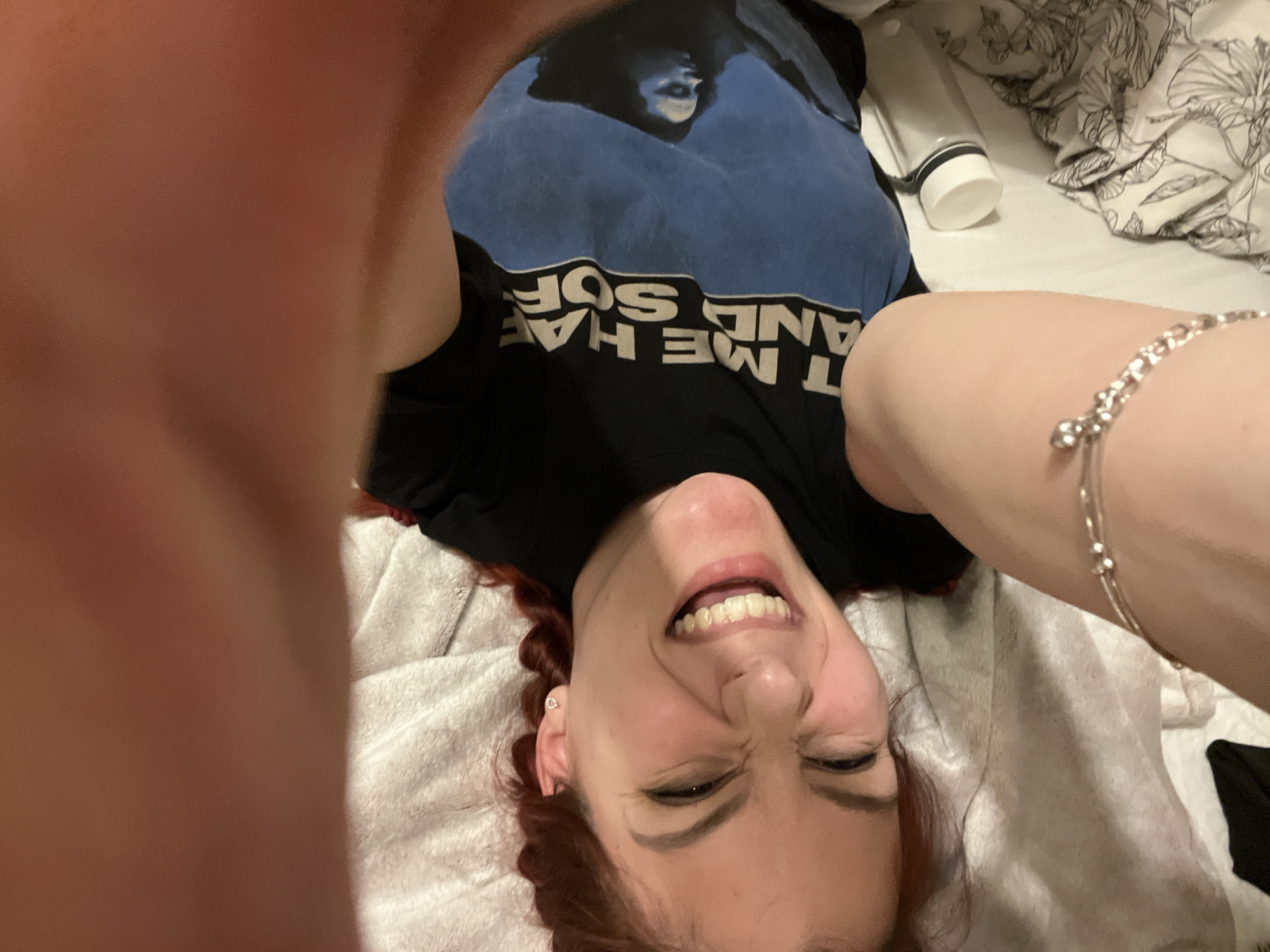 A young, light-skinned woman with red hair lies on a bed covered with a white blanket. She is wearing a black t-shirt with a blue design and white lettering. The photo appears to be a selfie taken from an unusual angle, showing her face upside down with a wide smile. Her eyes are squinted shut, and her arms are raised on either side of her head, partially obscuring the view. A silver bracelet is visible on one of her arms. In the background, there is a white water bottle and a patterned white sheet. The location cannot be identified as Sofia, Bulgaria based on the visual information provided.
