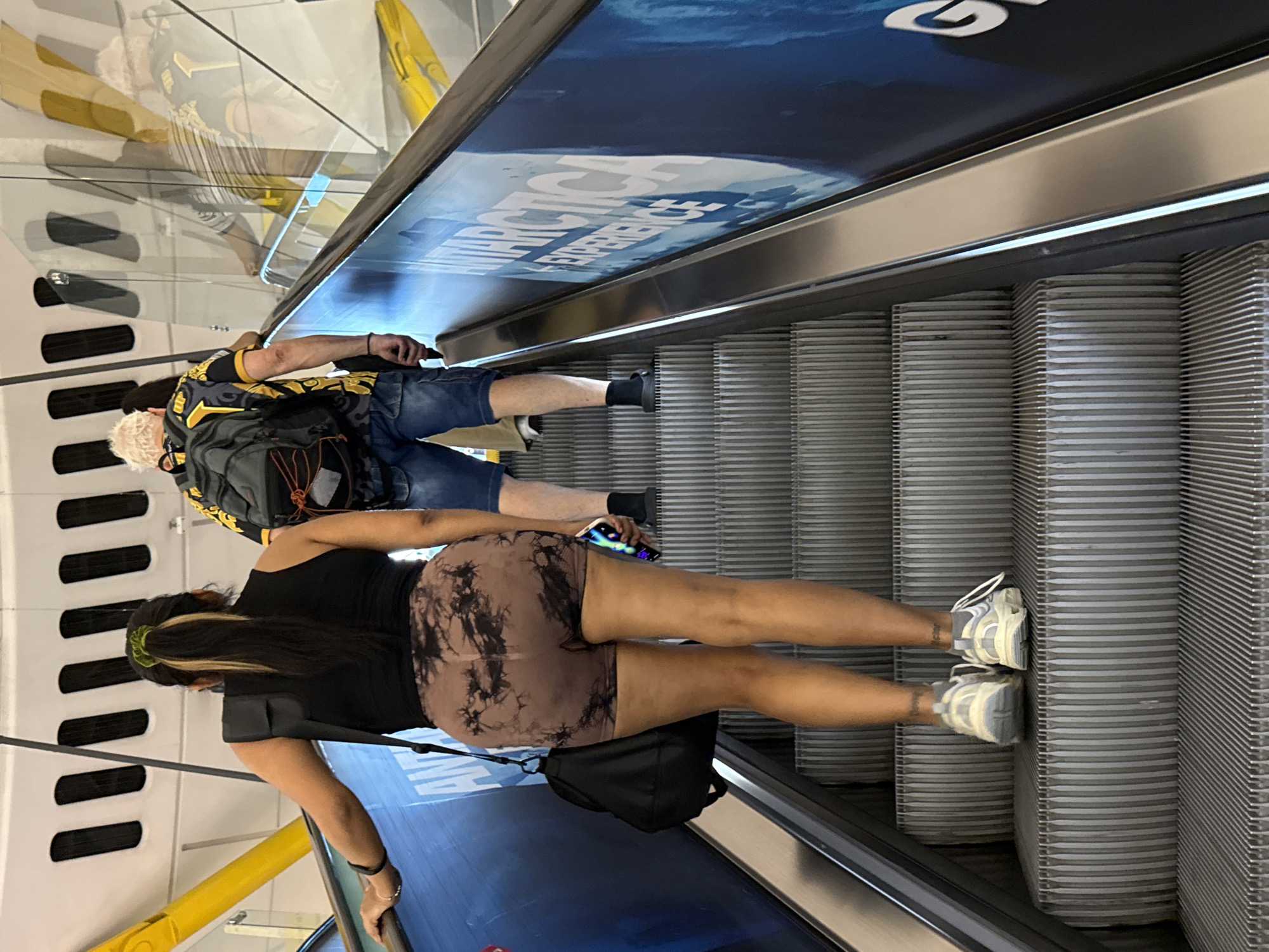 In a Barcelona setting, two individuals are seen ascending an escalator. The person in the foreground, viewed from the rear, is a woman with dark hair pulled back with a green scrunchie and streaks of blonde. She is wearing a black tank top and tie-dye shorts in shades of brown and black. On her right shoulder, a black crossbody bag is visible. She is holding onto the escalator handrail with her left hand. Her right hand holds a dark-colored smartphone. She is wearing white athletic shoes with gray accents.
Behind her, a man with short, white hair is also on the escalator. He is wearing a black and yellow patterned short-sleeved shirt, denim shorts, and black socks with dark shoes. He has a backpack on, with gray and orange detailing. He is holding the handrail with his right hand.
The escalator has metal steps with horizontal ridges. Above the escalator, a large advertisement is displayed, featuring images of ice and the text "ANTARCTICA EXPERIENCE" in white, bold lettering against a dark blue background. The scene appears to be indoors, possibly in a mall or airport, given the presence of escalators and large advertising. The lighting suggests it is daytime.