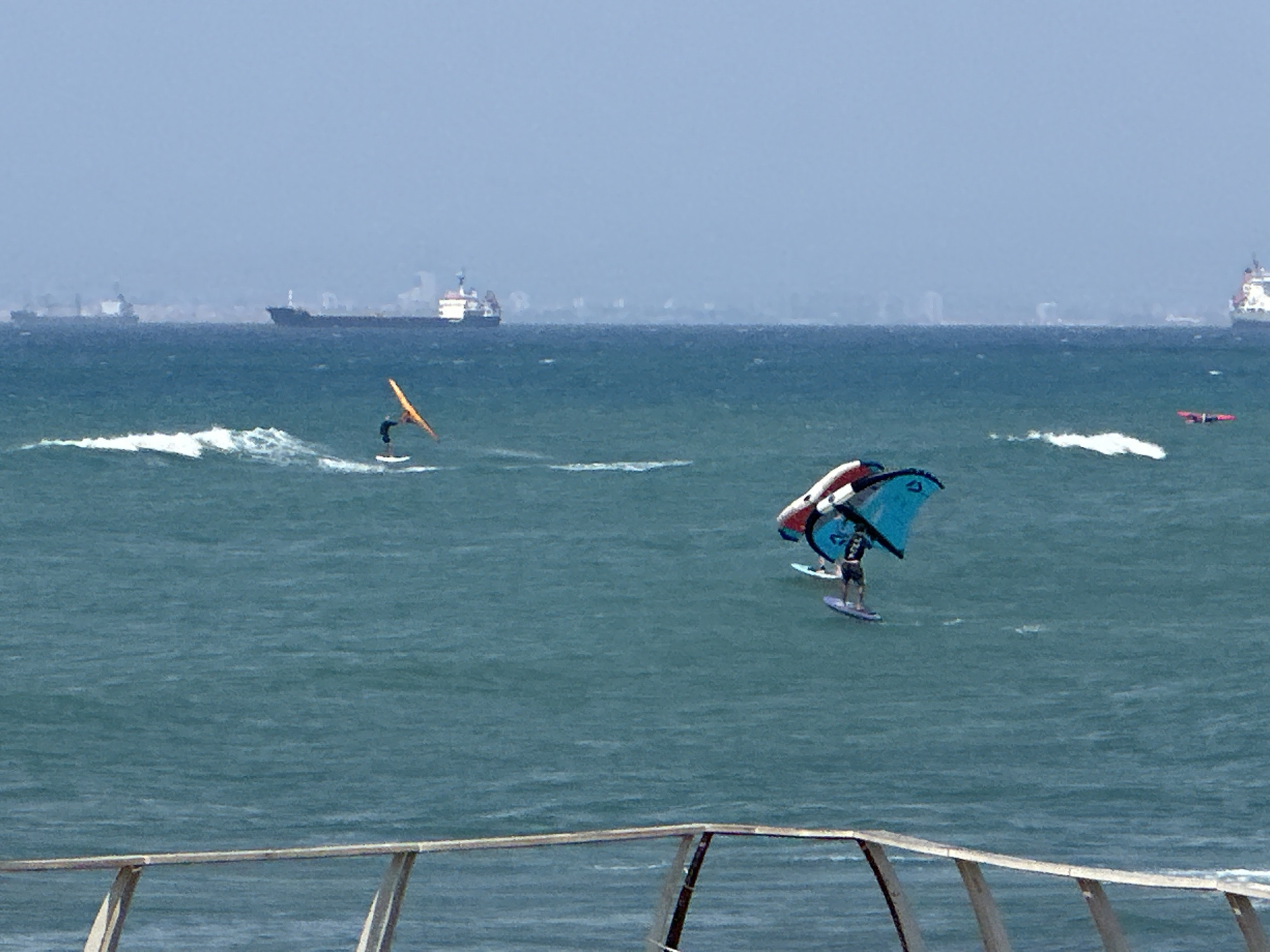 In the waters off Haifa, Israel, several individuals are engaged in wind-powered watersports. In the foreground, a person on a surfboard is using a large blue and white sail, appearing to be wing foiling. To the left, another person on a surfboard is also windsurfing, with an orange sail. In the background, several cargo ships are visible on the horizon, suggesting a busy port area. The water is a deep blue-green, with moderate waves. A railing structure is present in the extreme foreground.