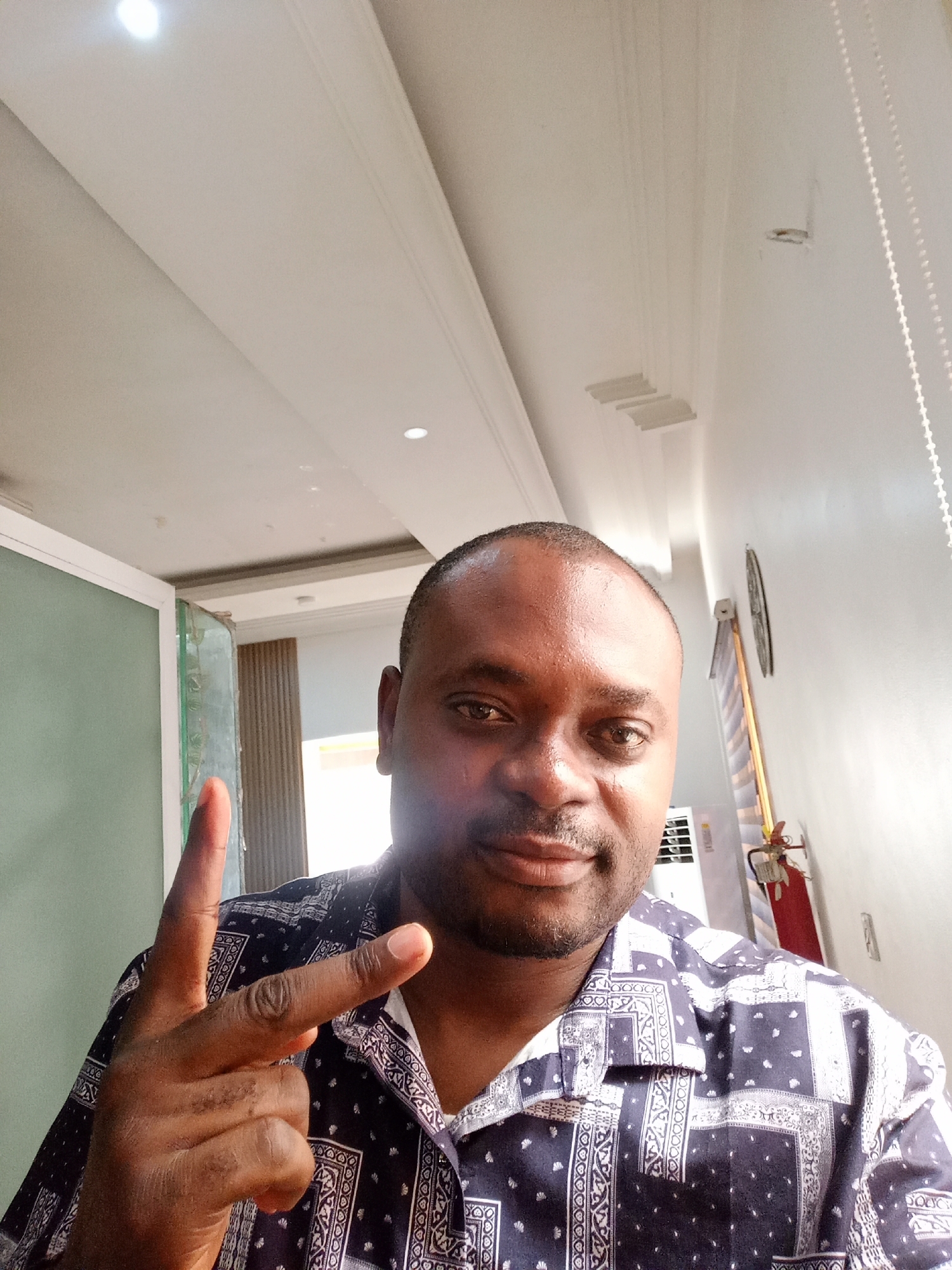 A man is taking a selfie indoors in Gwarinpa, Nigeria. The man, of African descent, is facing the camera and gesturing with his hand, holding up two fingers. He's wearing a navy blue button-down shirt with a white paisley pattern. Behind him, the room appears to have white walls and ceilings. A window or door is visible in the background, along with an air conditioner and a fire extinguisher mounted on the wall. A clock is also hanging on the wall. The lighting appears to be artificial and bright.