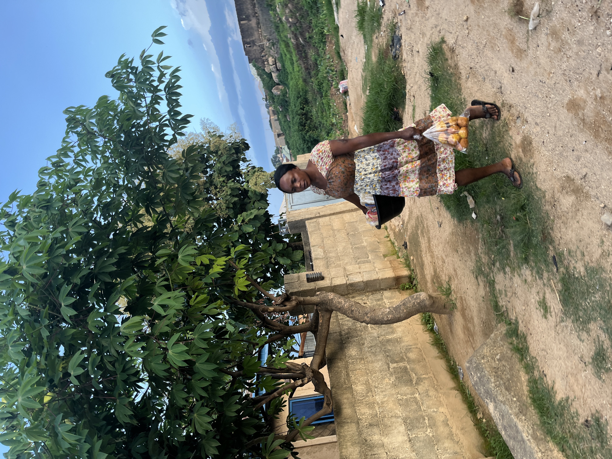 A woman, likely of African descent, stands outdoors under a clear blue sky with scattered clouds. She is wearing a floral print dress and flip-flops. In her right hand, she holds a clear plastic bag filled with round, orange-colored fruits, possibly oranges or tangerines. Her left arm is bent at the elbow, and she is holding a dark-colored bucket or bowl.
The setting appears to be a dusty, unpaved area with patches of grass. In the background, there is a brick building and a sloping, grassy hill. A large tree with green leaves is prominent in the foreground, on the left side of the image. The overall lighting suggests it is daytime, likely late afternoon given the angle of the sunlight and the warm tones. The woman is looking towards the camera and appears to be smiling slightly. The scene evokes a sense of everyday life in a rural or semi-rural environment. There is no visible text in the image.